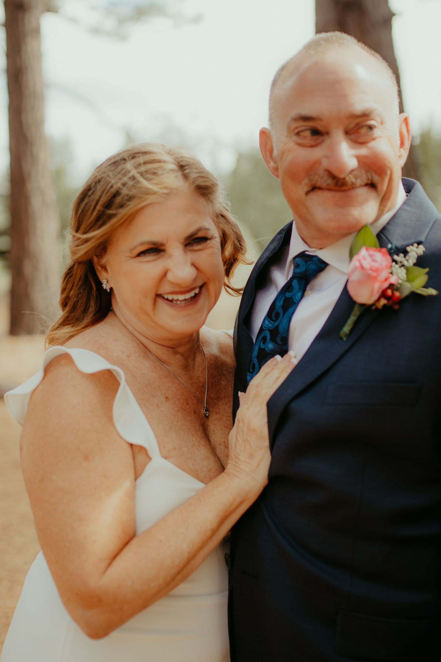 Happy mature bride and groom smile together in navy suit with pink boutonniere at outdoor wedding.