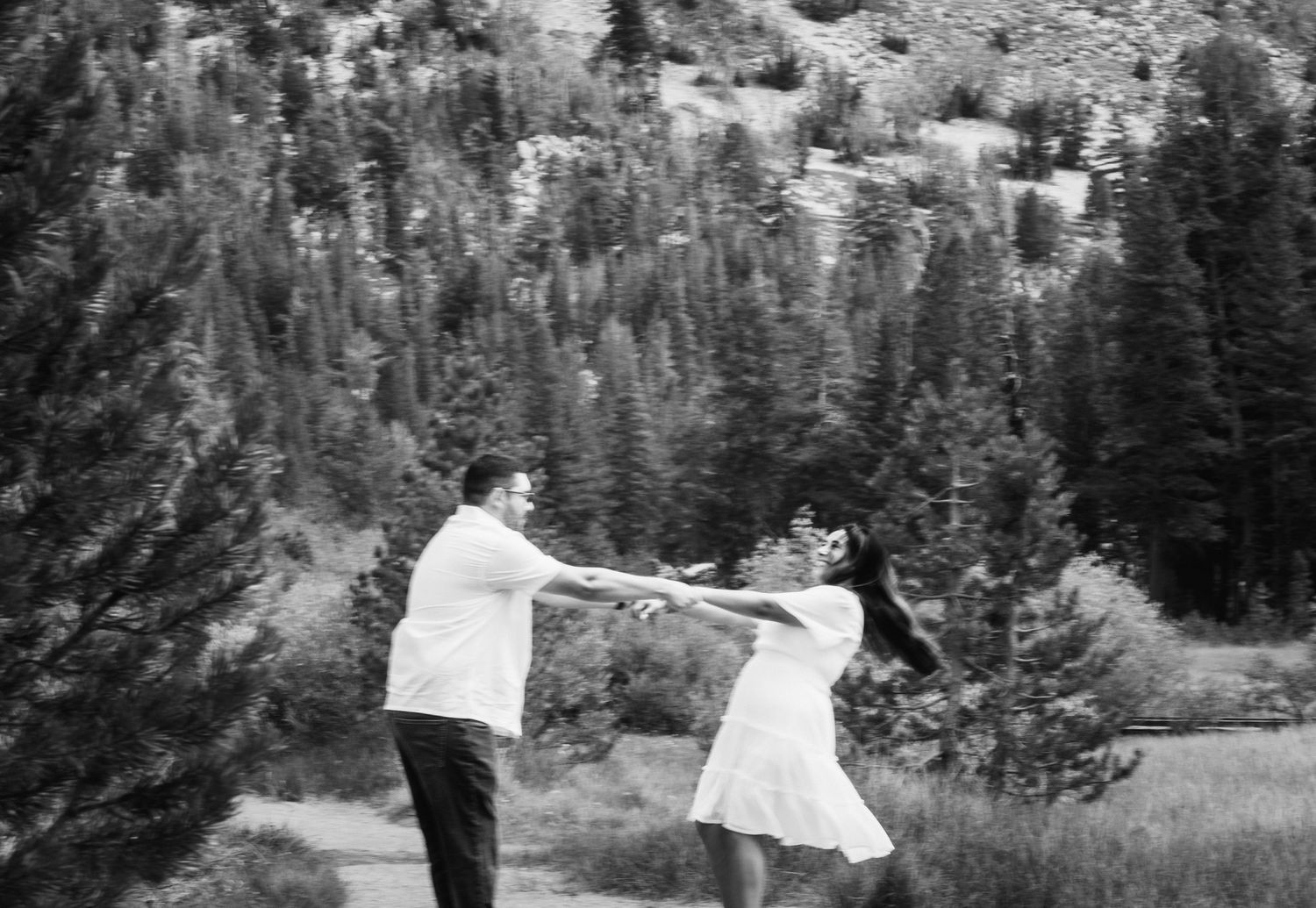 Black and white photo of a couple dancing and holding hands in a mountain meadow surrounded by pine trees.