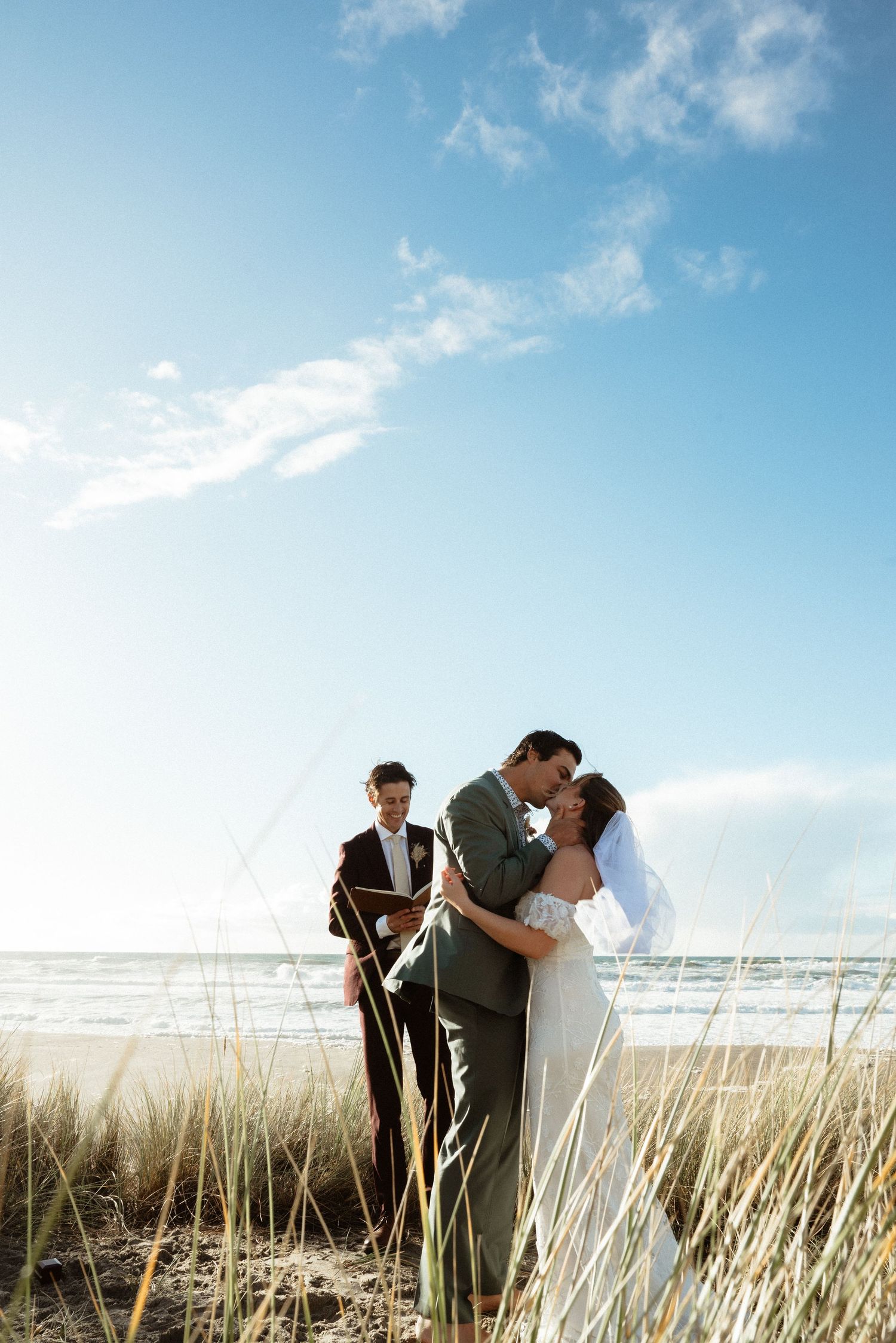 Bride and groom share a kiss during their beachside wedding ceremony as the officiant looks on beneath a blue sky with dune grass.