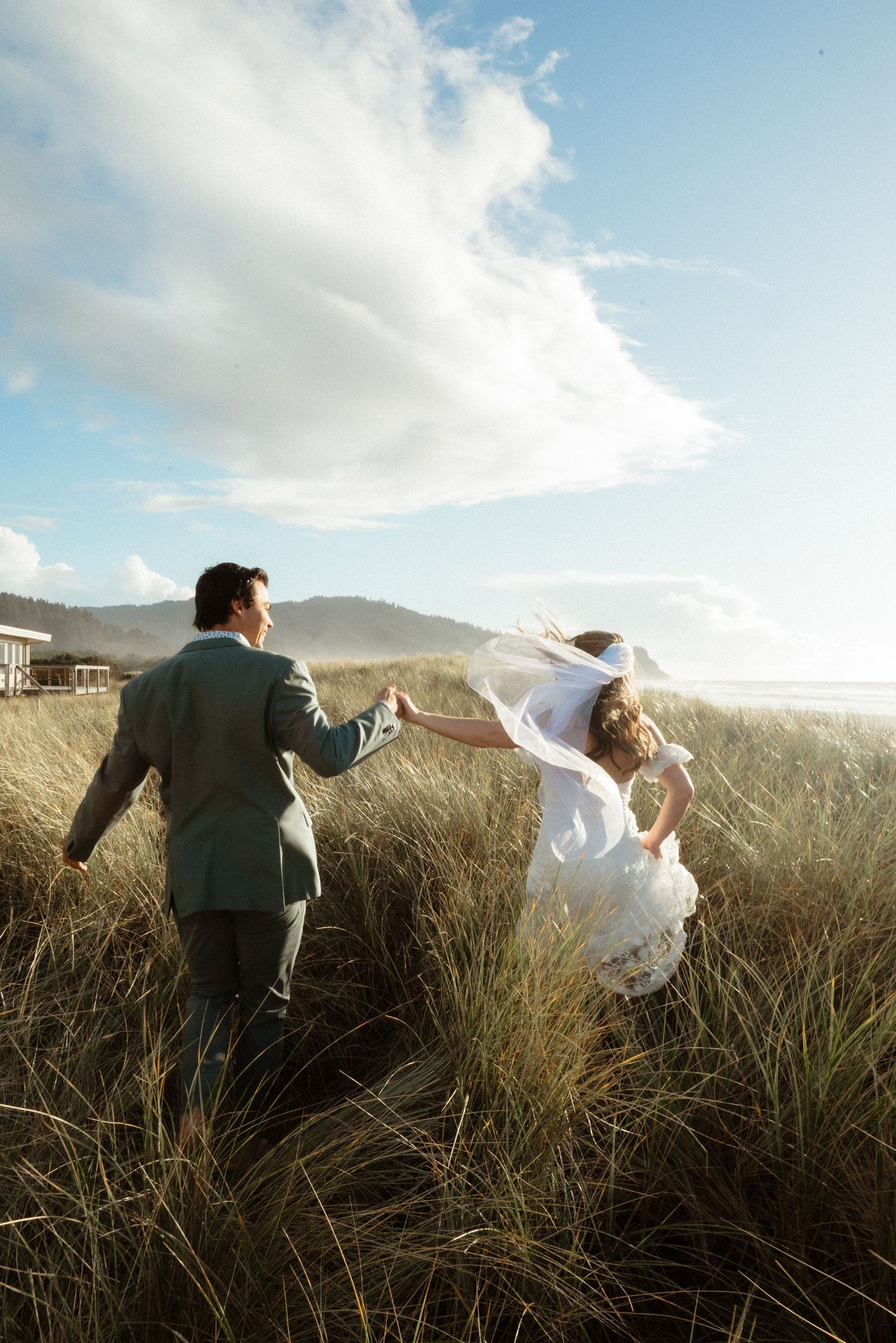 Bride and groom hold hands and walk through windswept dune grass toward the ocean as her veil blows in the breeze.