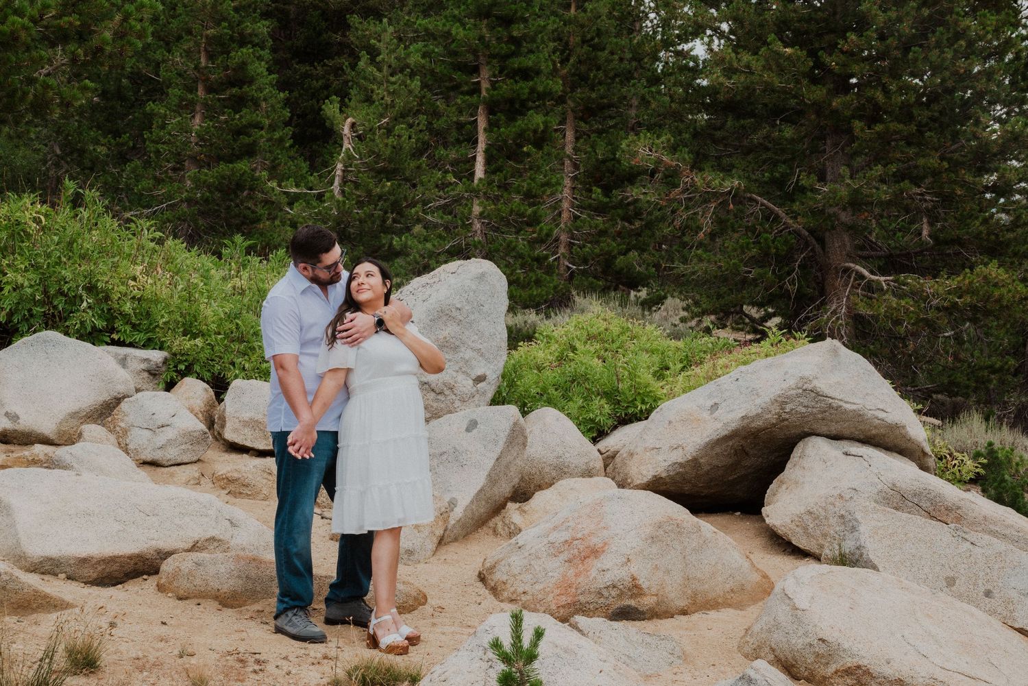 Couple embracing among large boulders surrounded by pine trees in a rocky mountain landscape.