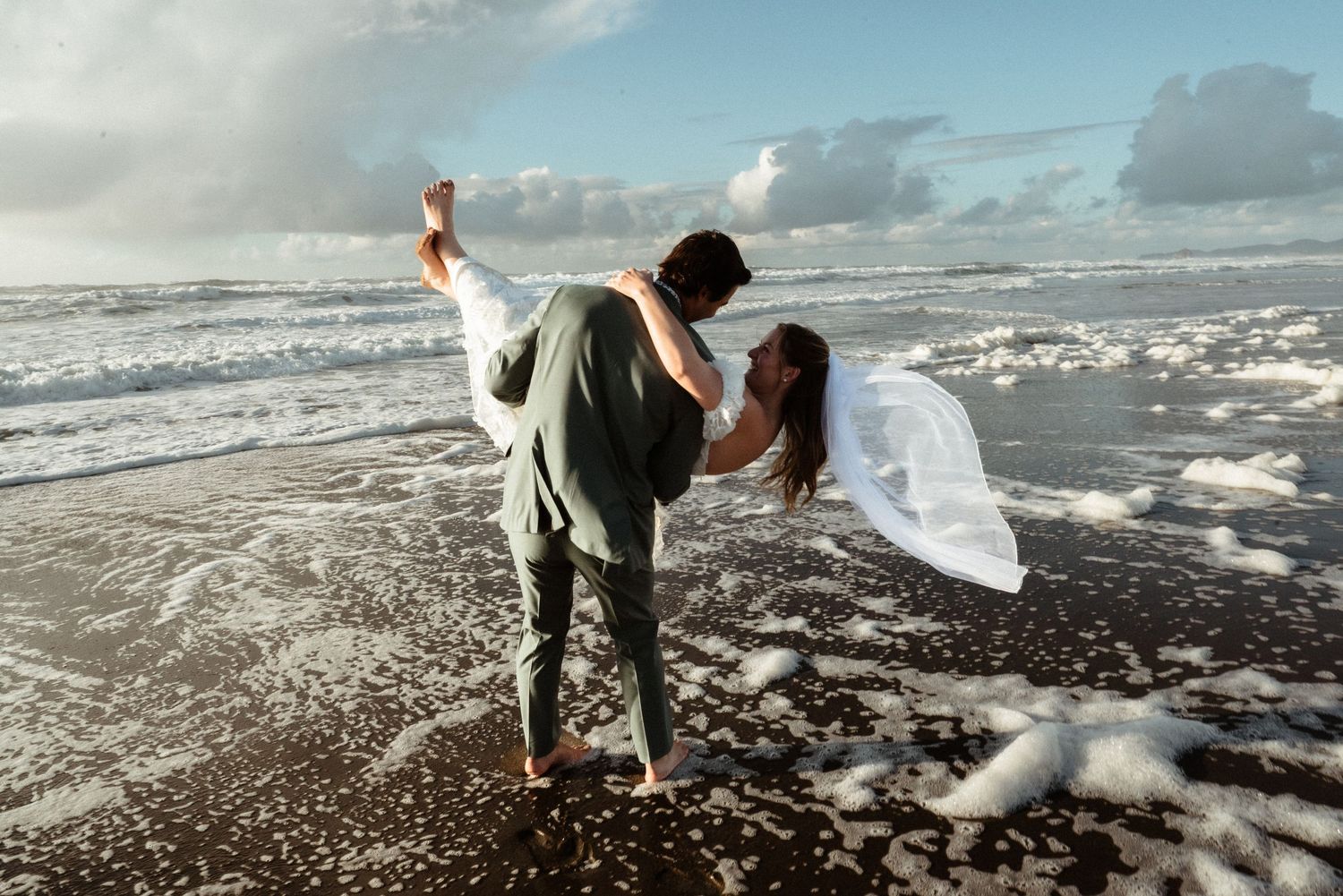 Couple in wedding attire dancing romantically on a black sand beach with ocean waves and foam in the background.