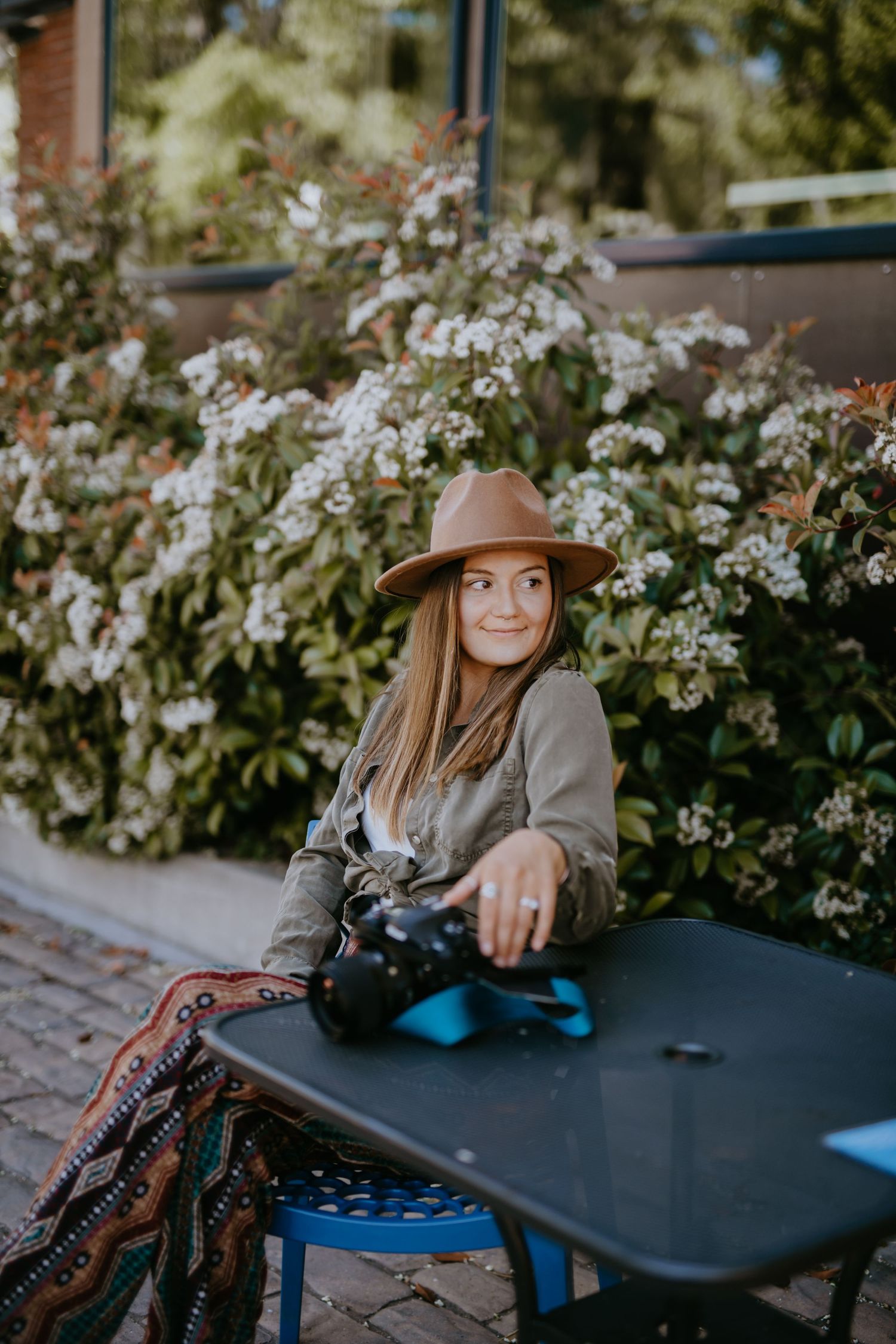 Smiling woman in brown hat holds camera while sitting at outdoor cafe table.