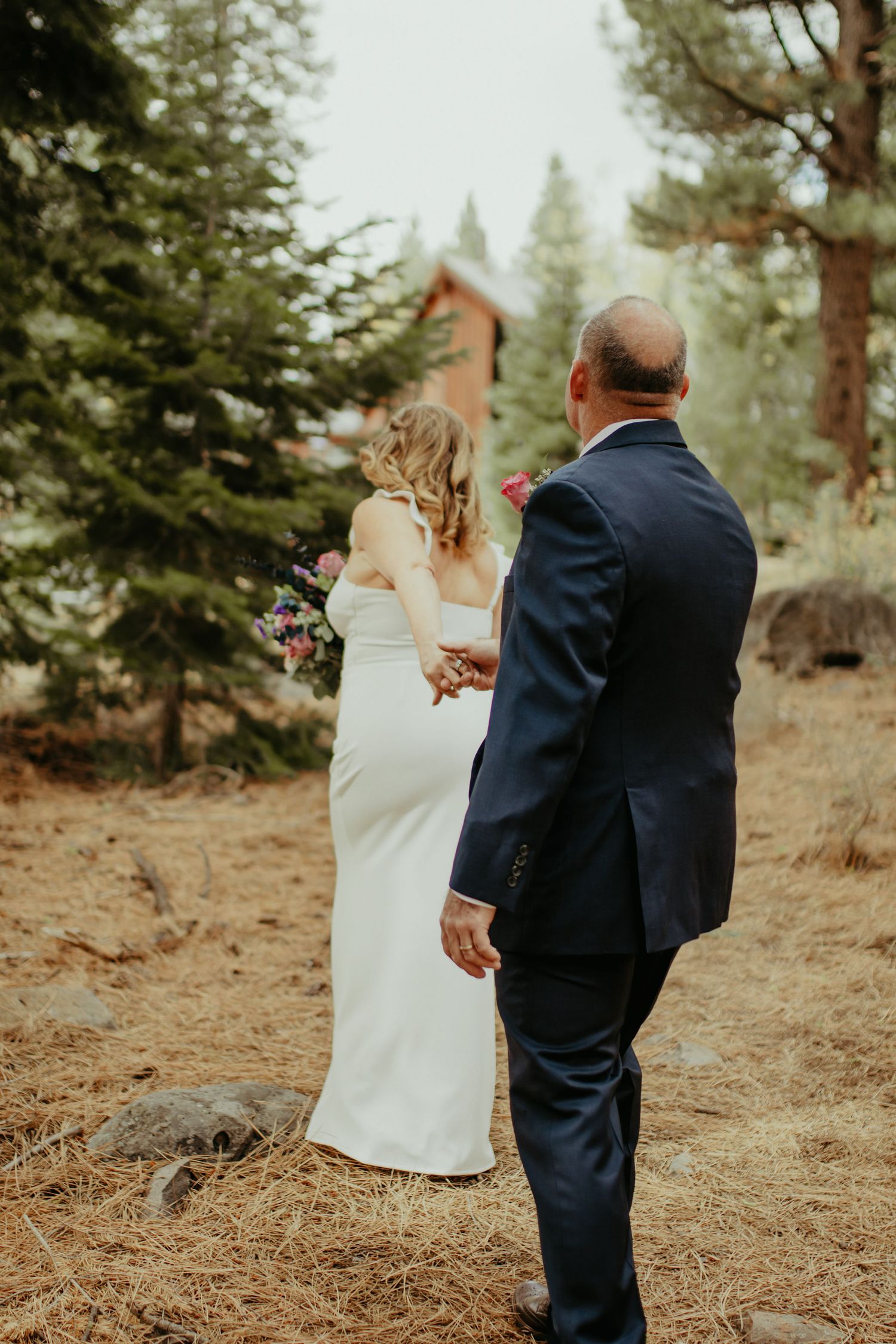 Bride in white gown holding bouquet walks hand-in-hand with groom in navy suit through pine forest.