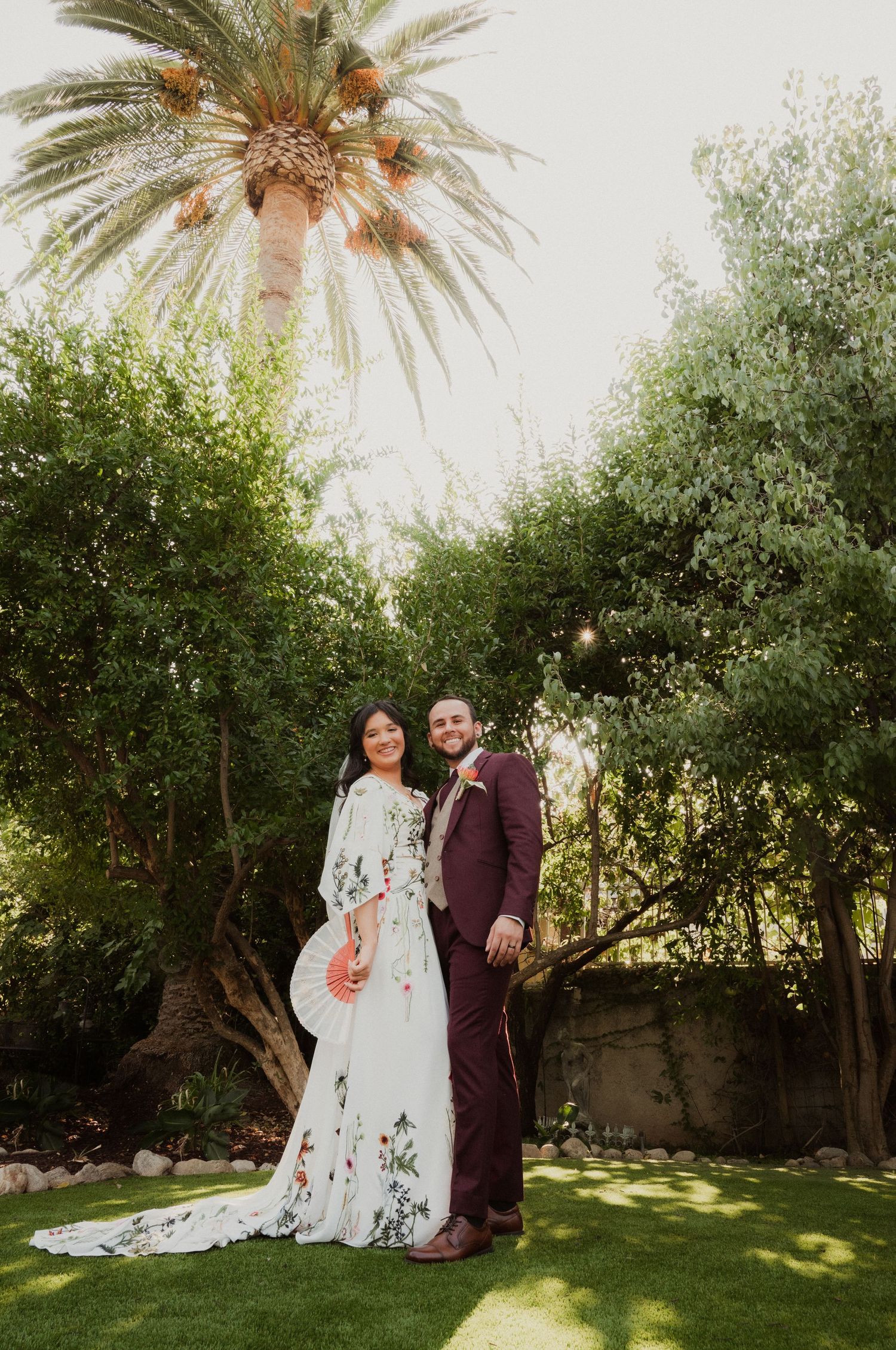 A bride in a floral gown and groom in burgundy suit pose together on green lawn beneath a tall palm tree.