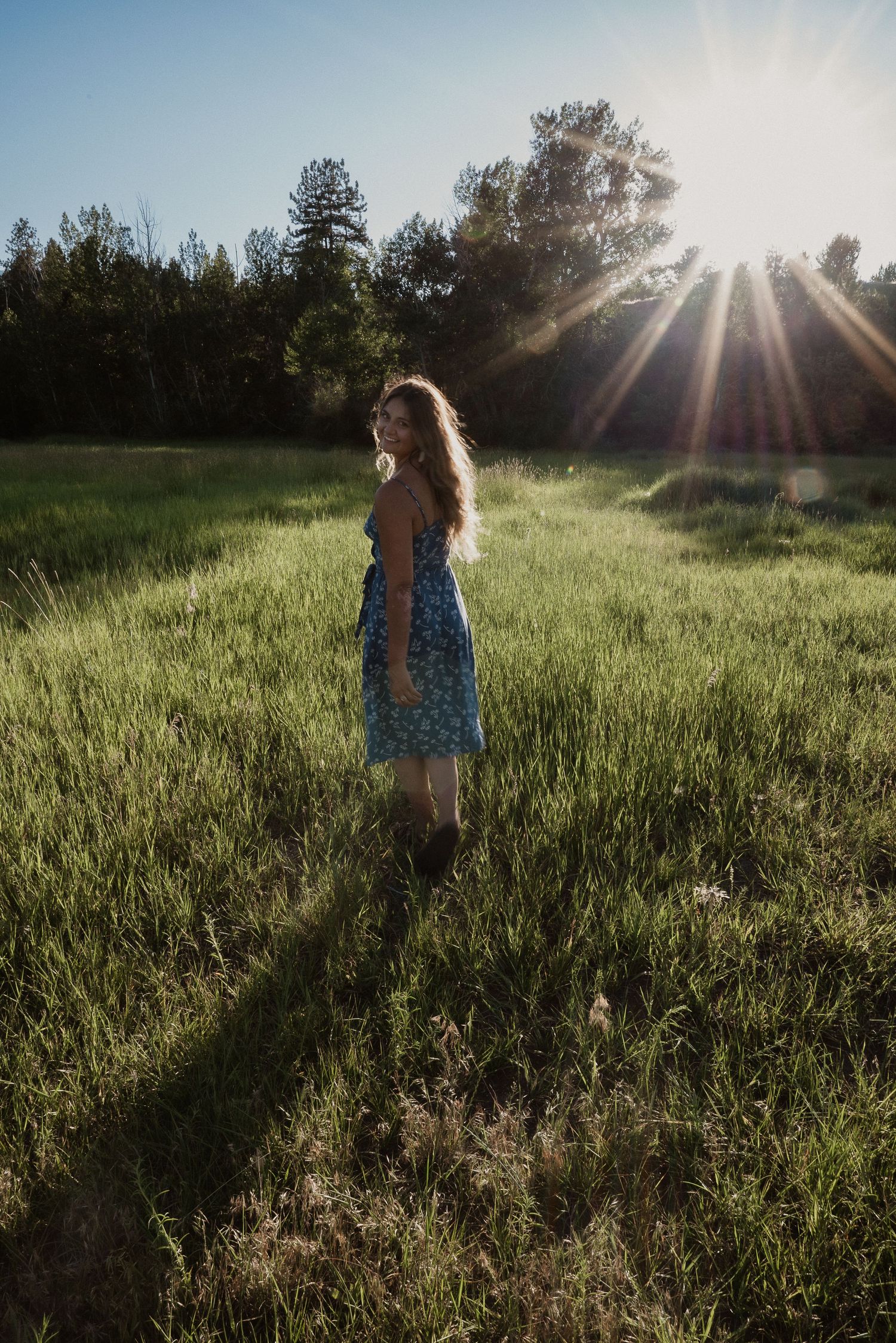 Woman in blue floral dress stands in sunlit green meadow with sun rays behind her.