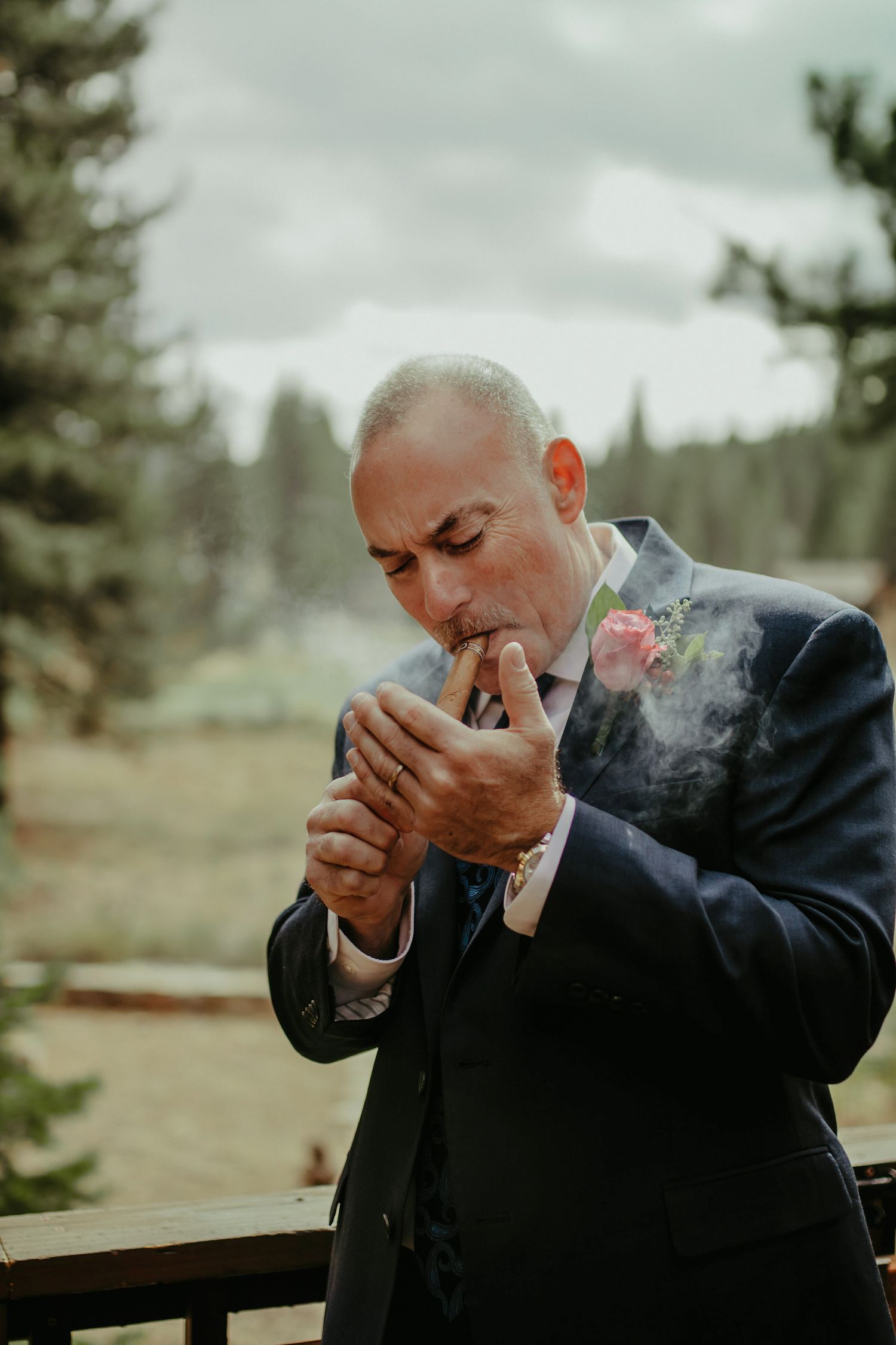 A groom in a navy suit with a pink boutonniere lights a cigar outdoors at a wedding venue surrounded by pine trees.