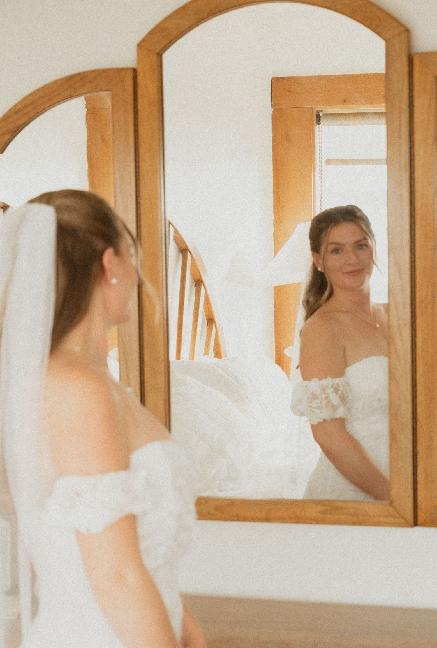 Bride in an off-the-shoulder lace wedding dress and veil admires her reflection in a wooden-framed mirror in a bright room.