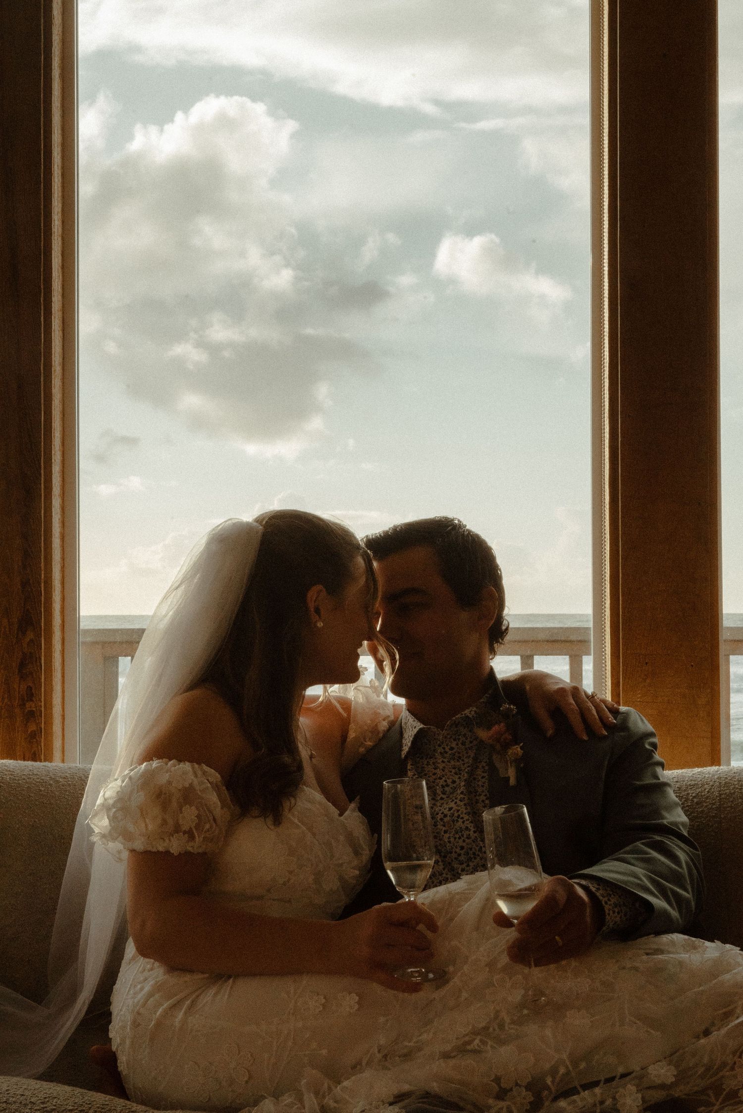 Bride in a lace wedding gown and veil shares an intimate moment with her groom, holding champagne glasses by a large window with cloudy sky.