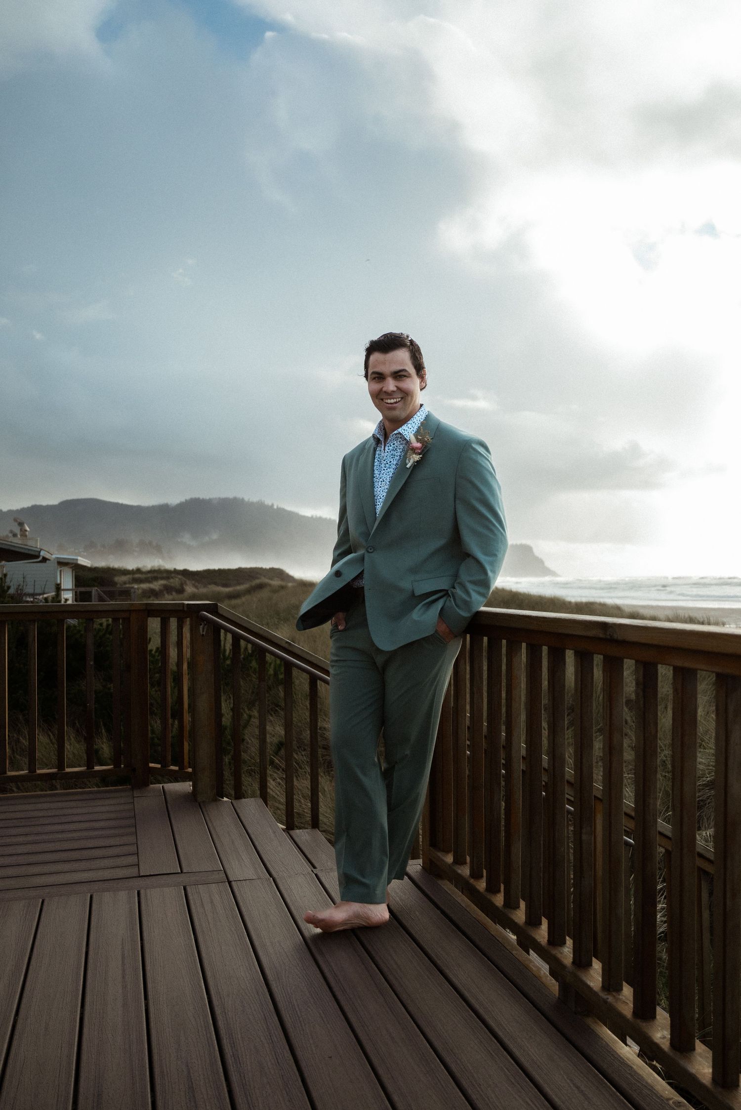 Smiling barefoot groom in a green suit with a boutonniere stands on a wooden deck overlooking a windswept coastal beach.