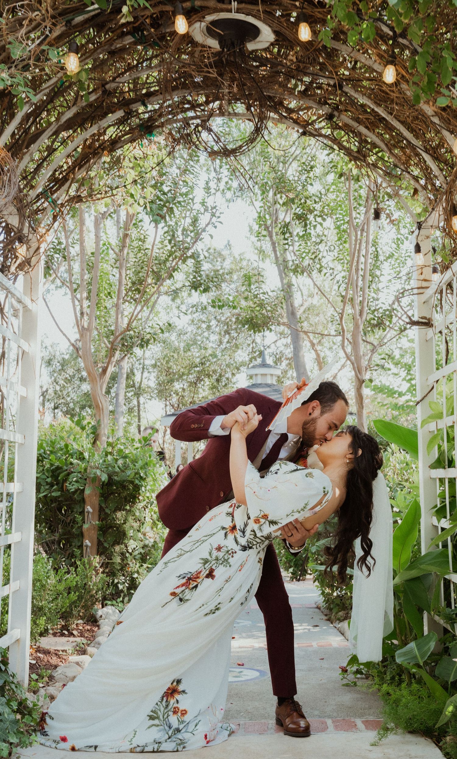 Bride and groom share a romantic dip kiss under a vine-covered arch with string lights at an outdoor garden wedding.