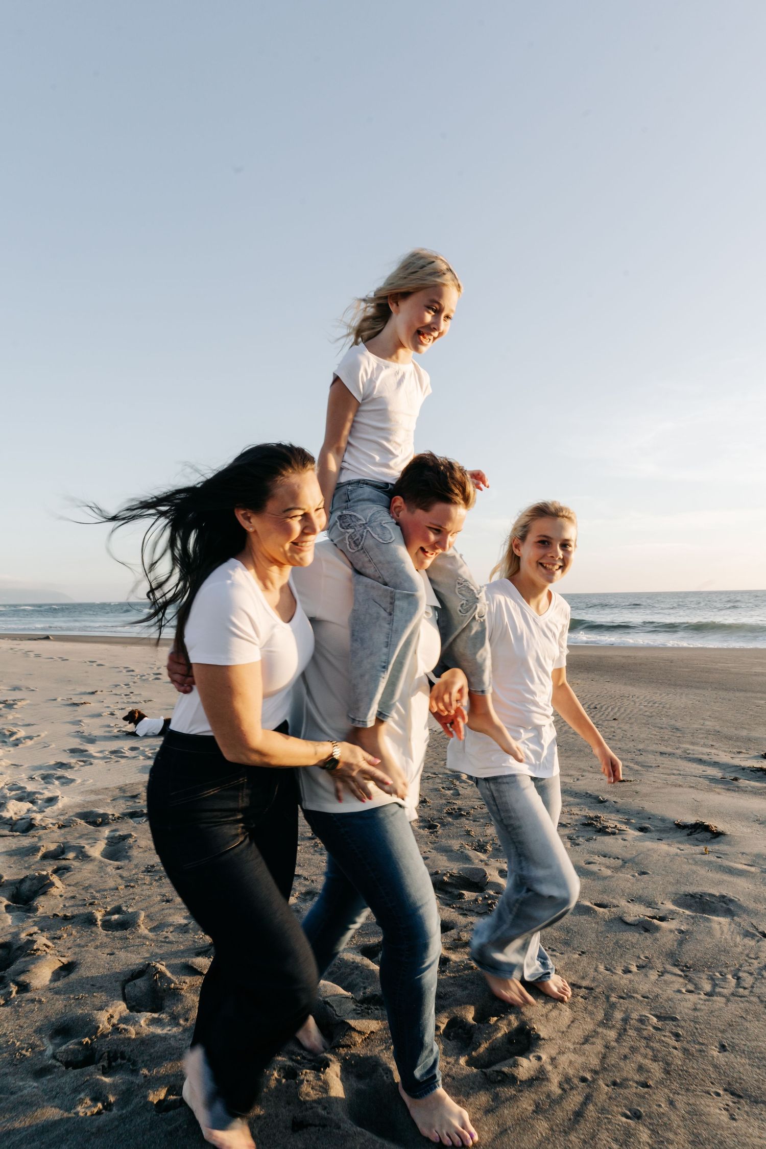 Happy family walking on beach at sunset, parents carrying child on shoulders, all wearing white shirts and jeans.