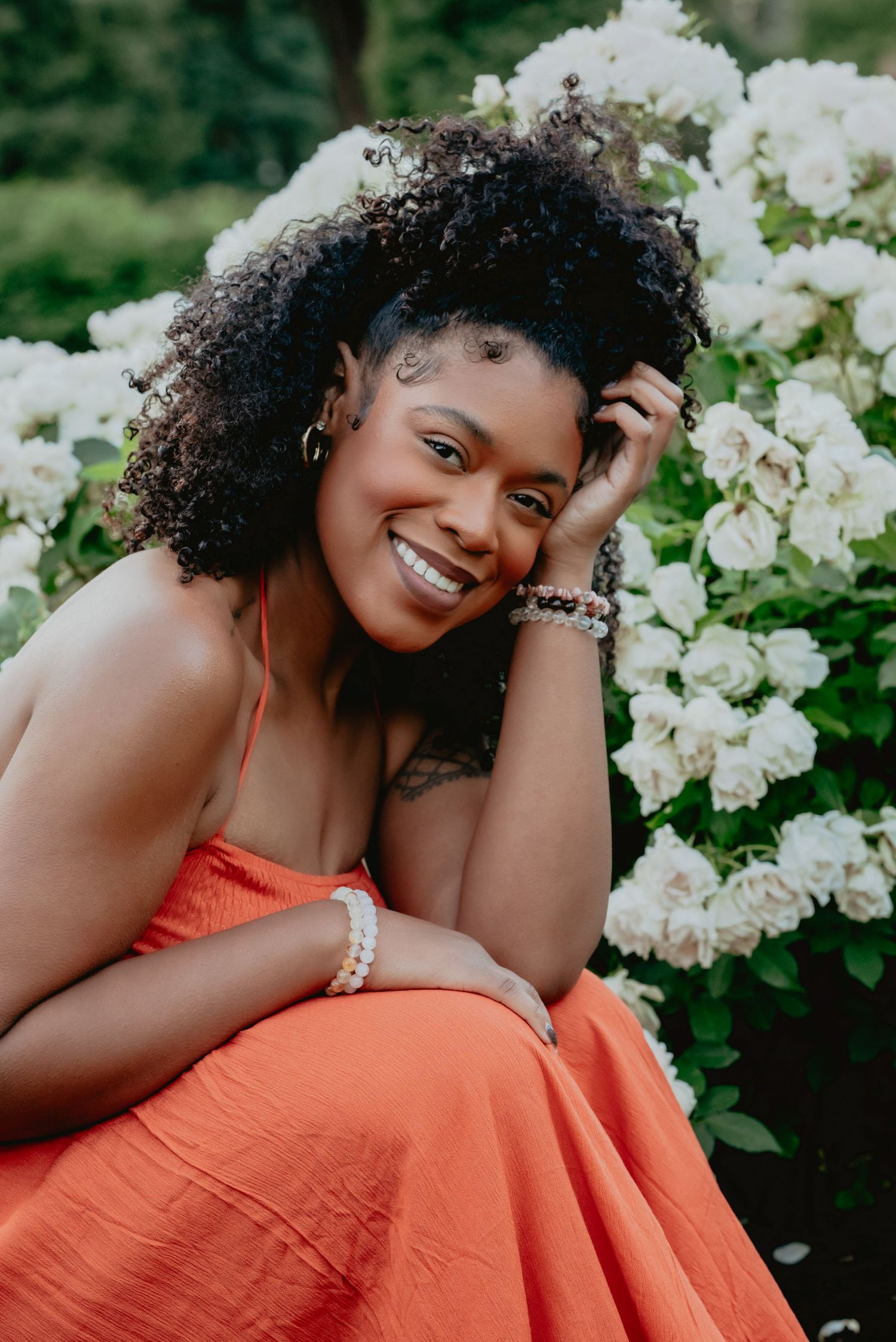 Smiling woman in orange dress with natural curly hair poses among white roses in a lush garden.