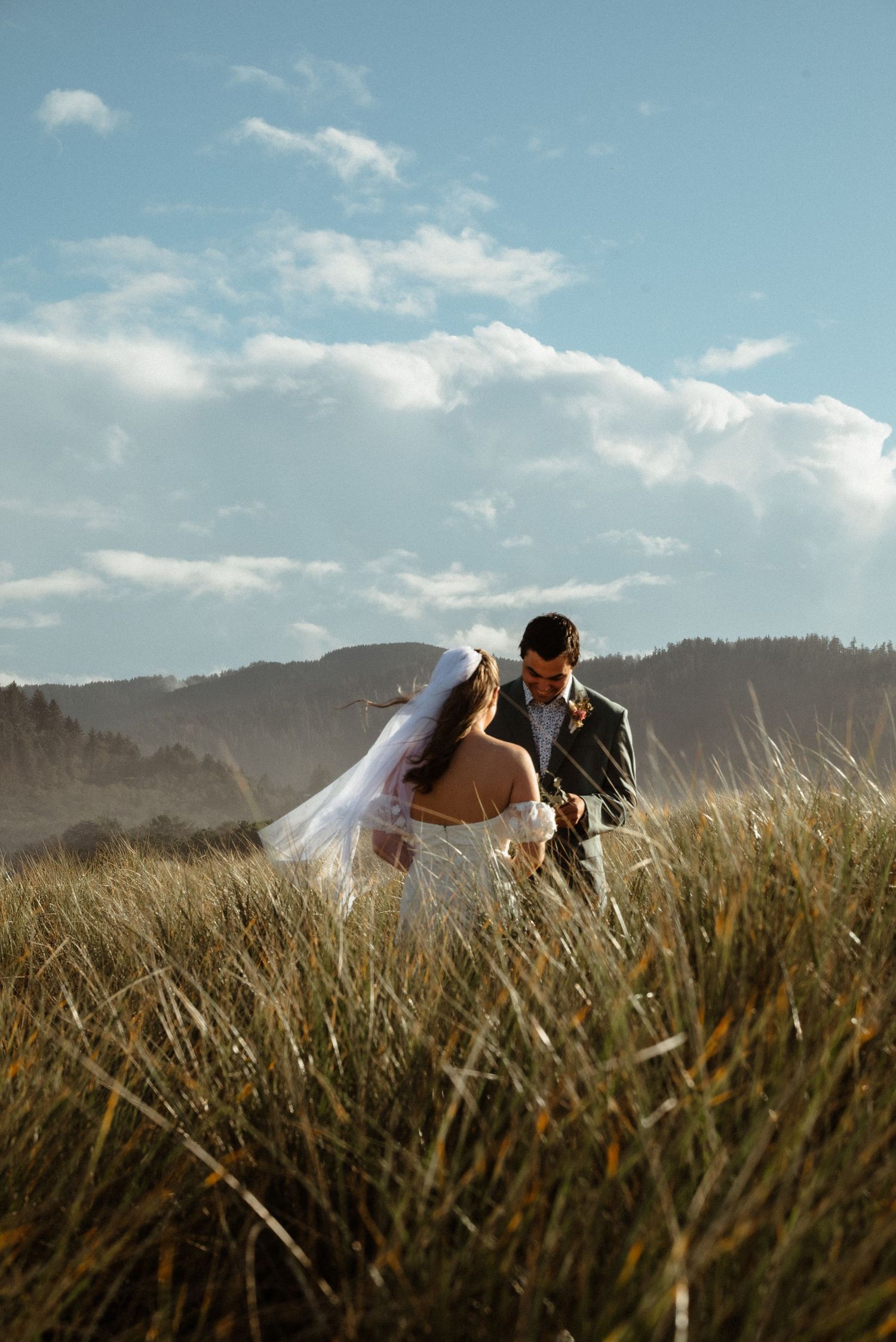 Bride in a long veil and groom in a gray suit share a quiet moment in tall golden grass beneath a blue sky.