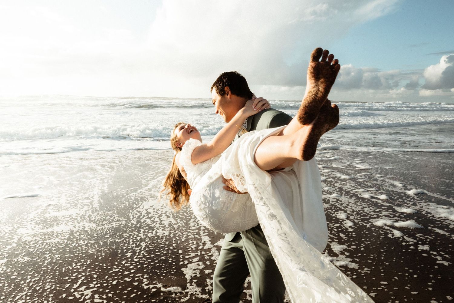 Groom carrying his barefoot bride in a white wedding dress along the wet sand of a sunlit ocean beach.