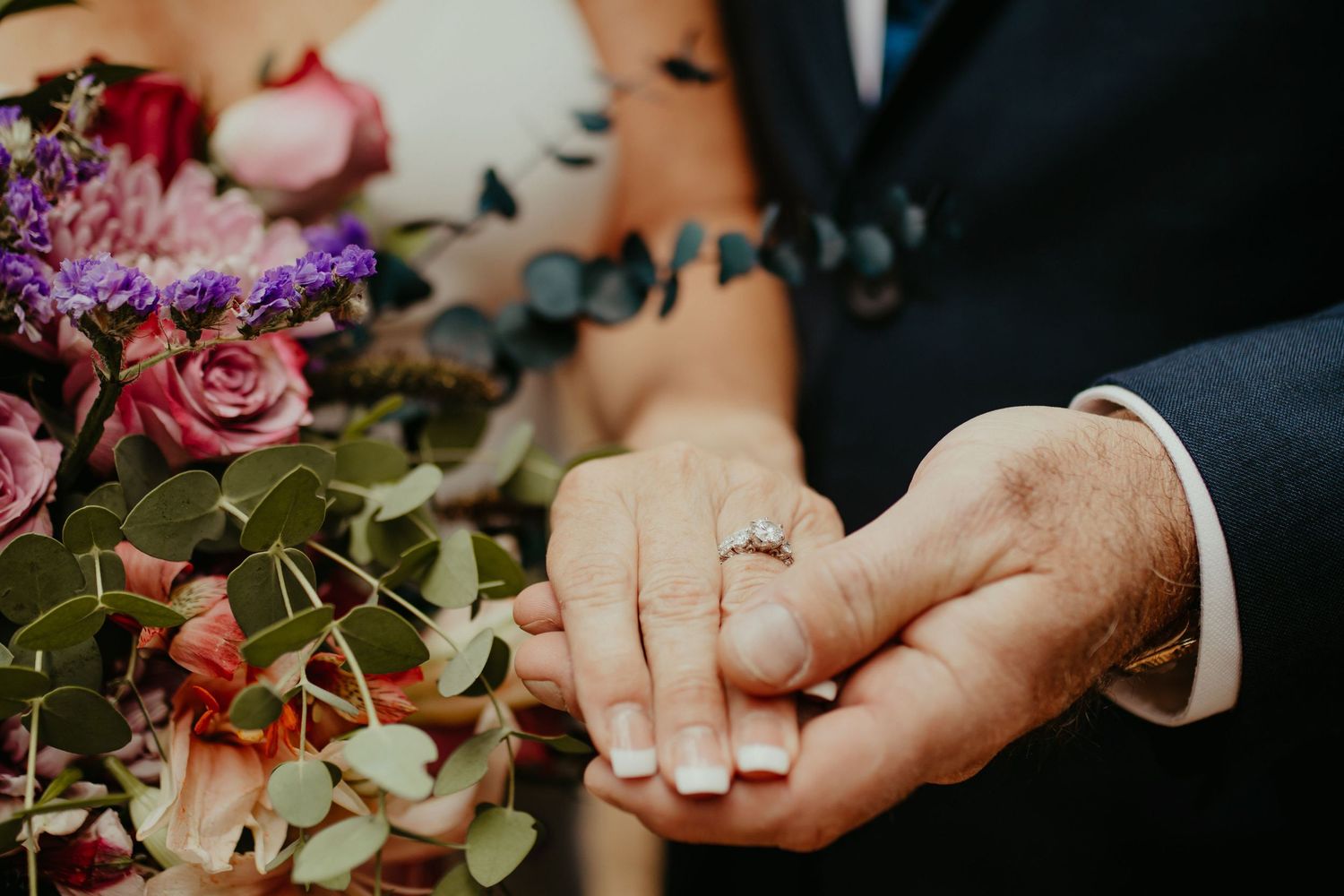 Bride and groom holding hands showing diamond engagement ring beside colorful wedding bouquet with roses and eucalyptus.