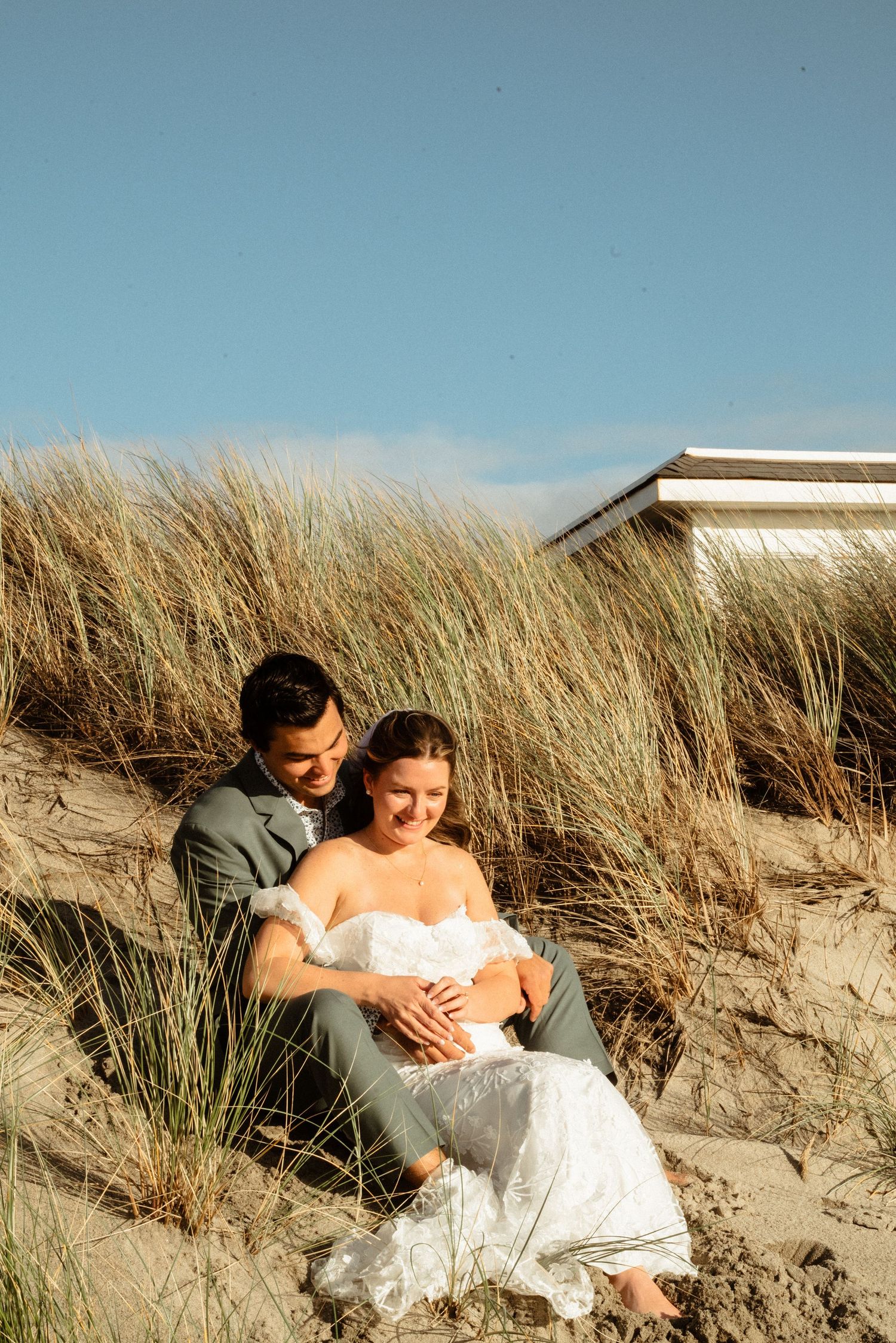 Newlywed couple in wedding attire sit close together among beach dune grass under a clear blue sky.