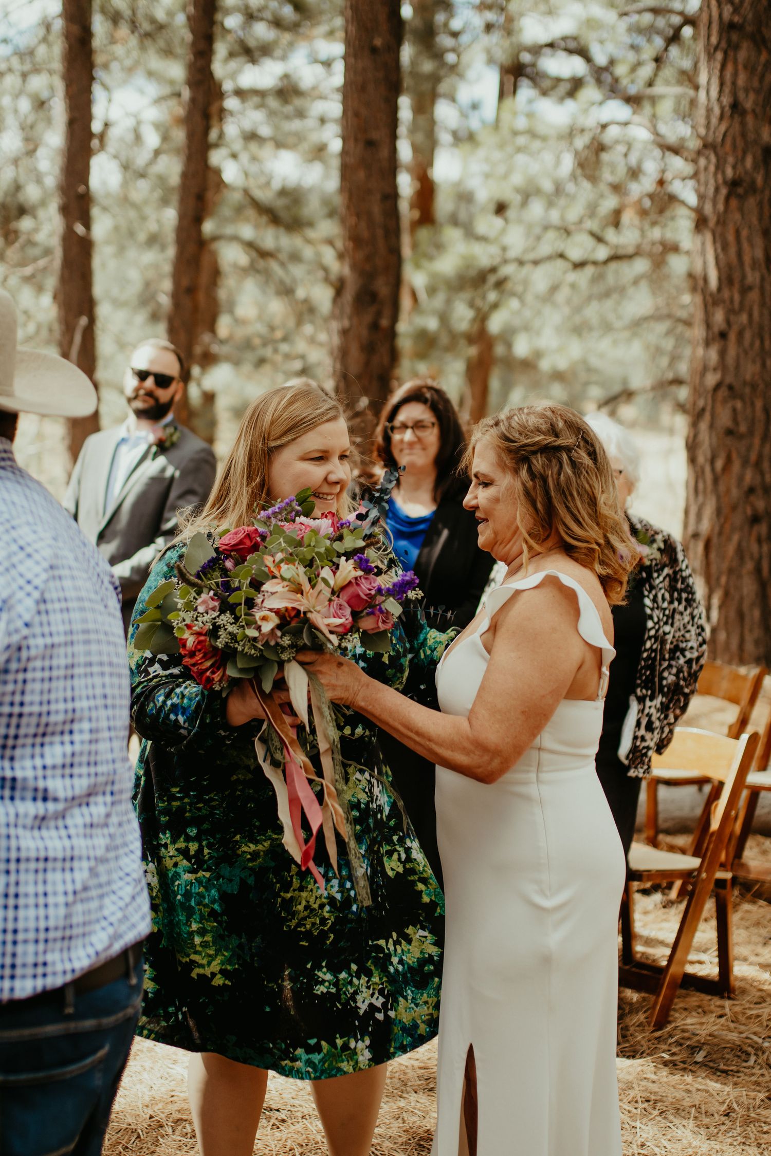 Bride in white dress receives bouquet from guest in floral dress at outdoor forest wedding ceremony.