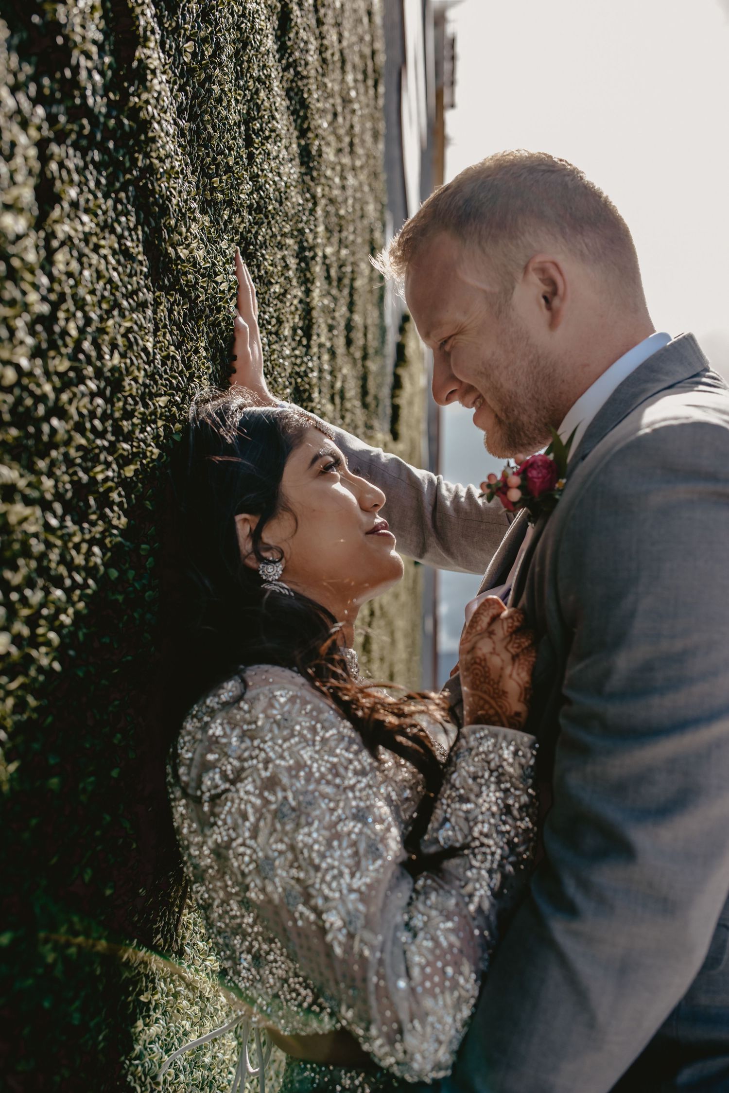 Interracial wedding couple sharing romantic moment against lush green ivy wall in sunlight.