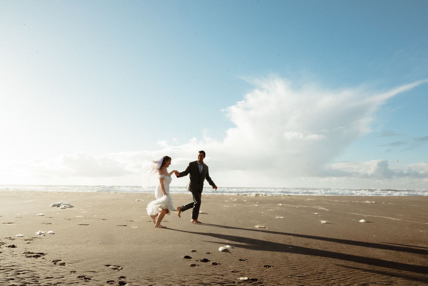 Bride in a white gown and groom in a dark suit hold hands and run along a wide sandy beach under a bright blue sky with billowing clouds.