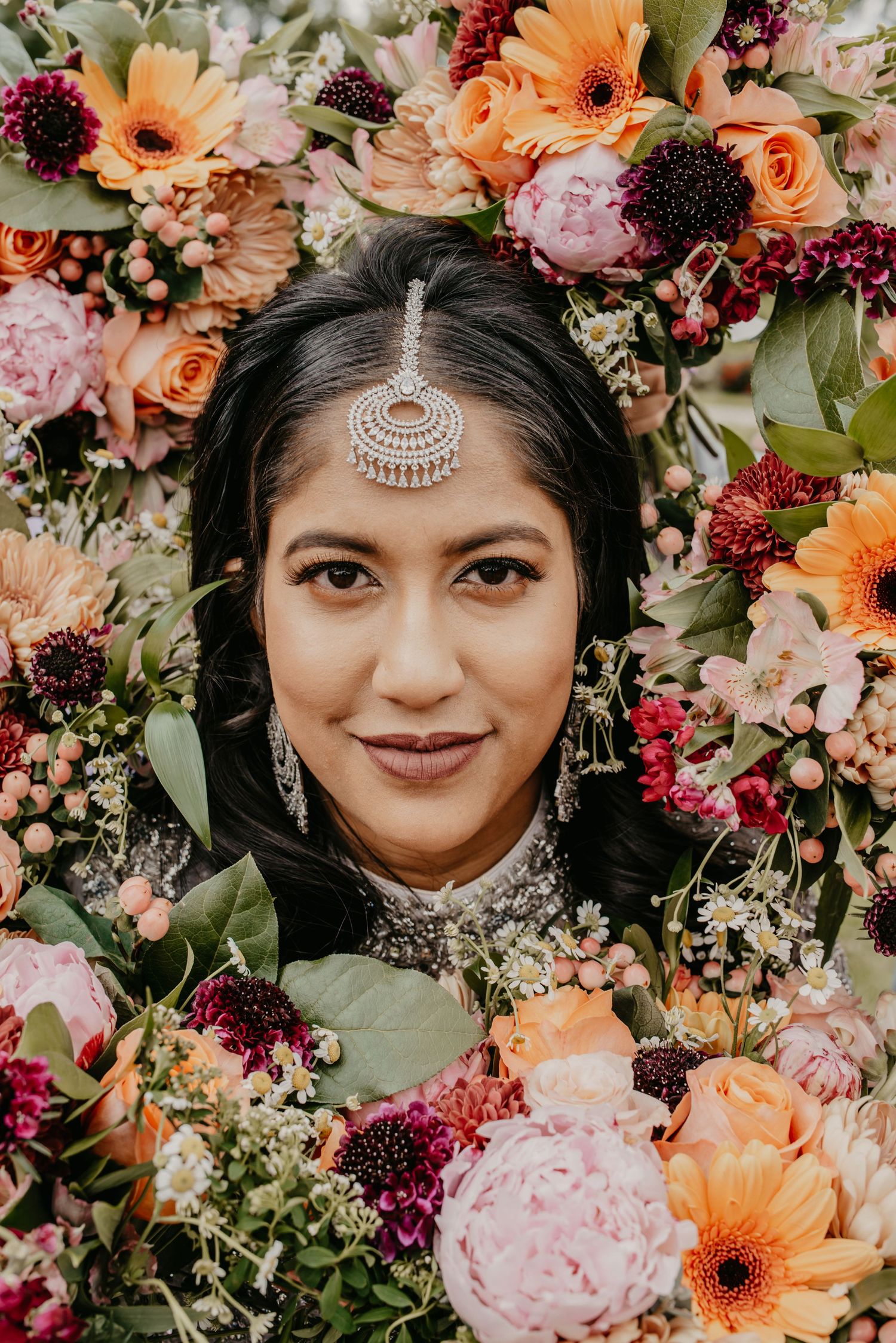 Indian bride surrounded by vibrant orange, pink, and burgundy flowers wearing traditional maang tikka jewelry.