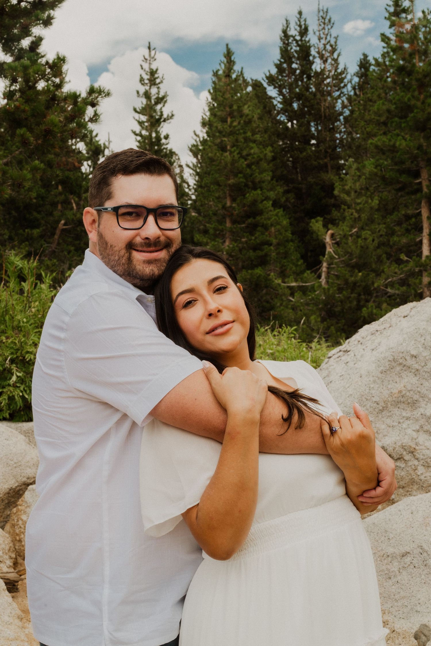 Couple embracing outdoors in white outfits with pine trees and rocky landscape in background.