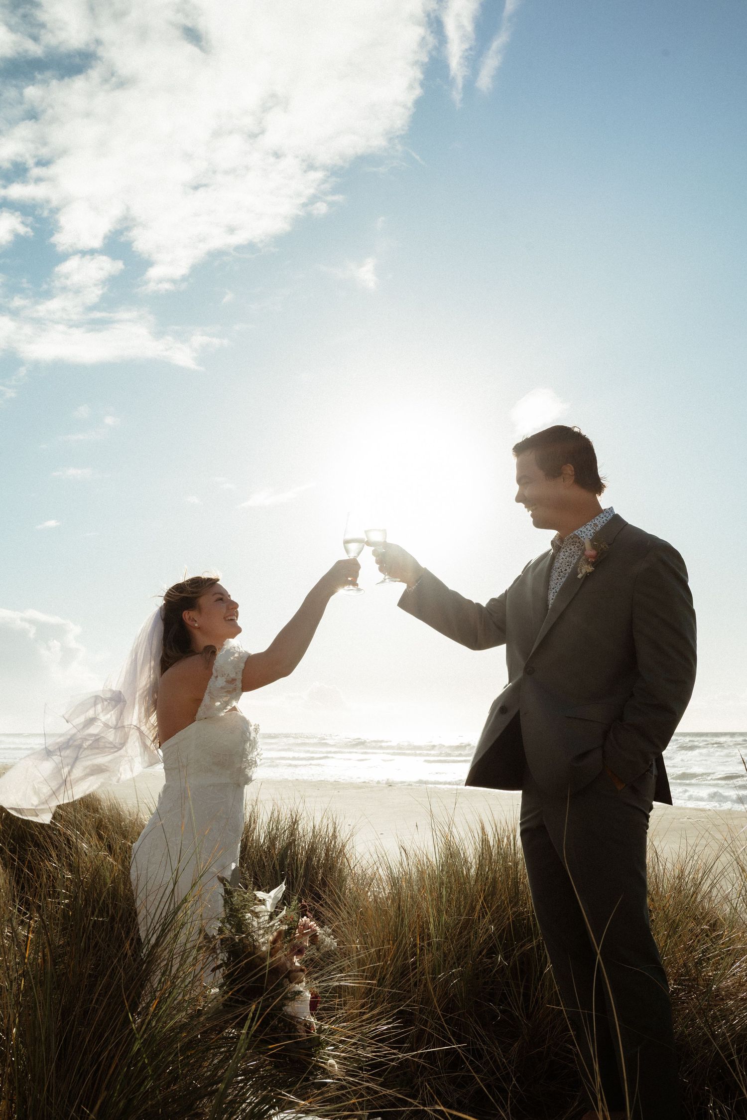 Bride and groom raise champagne glasses in a toast against a bright sun and ocean backdrop on the dunes.