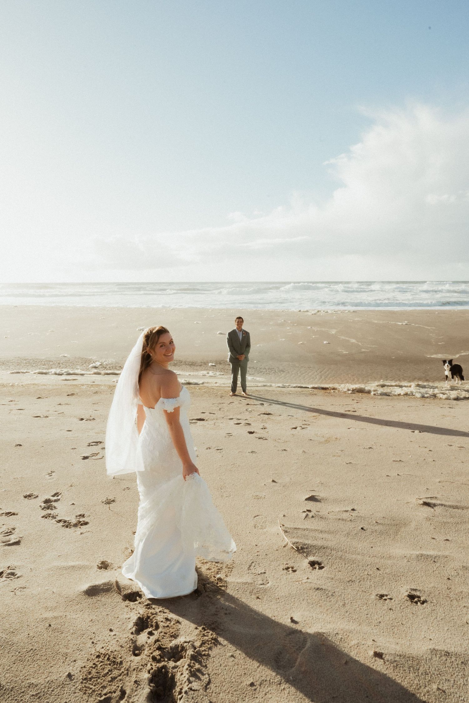 Bride in white off-shoulder gown and veil poses on sandy beach with groom and dog in background.