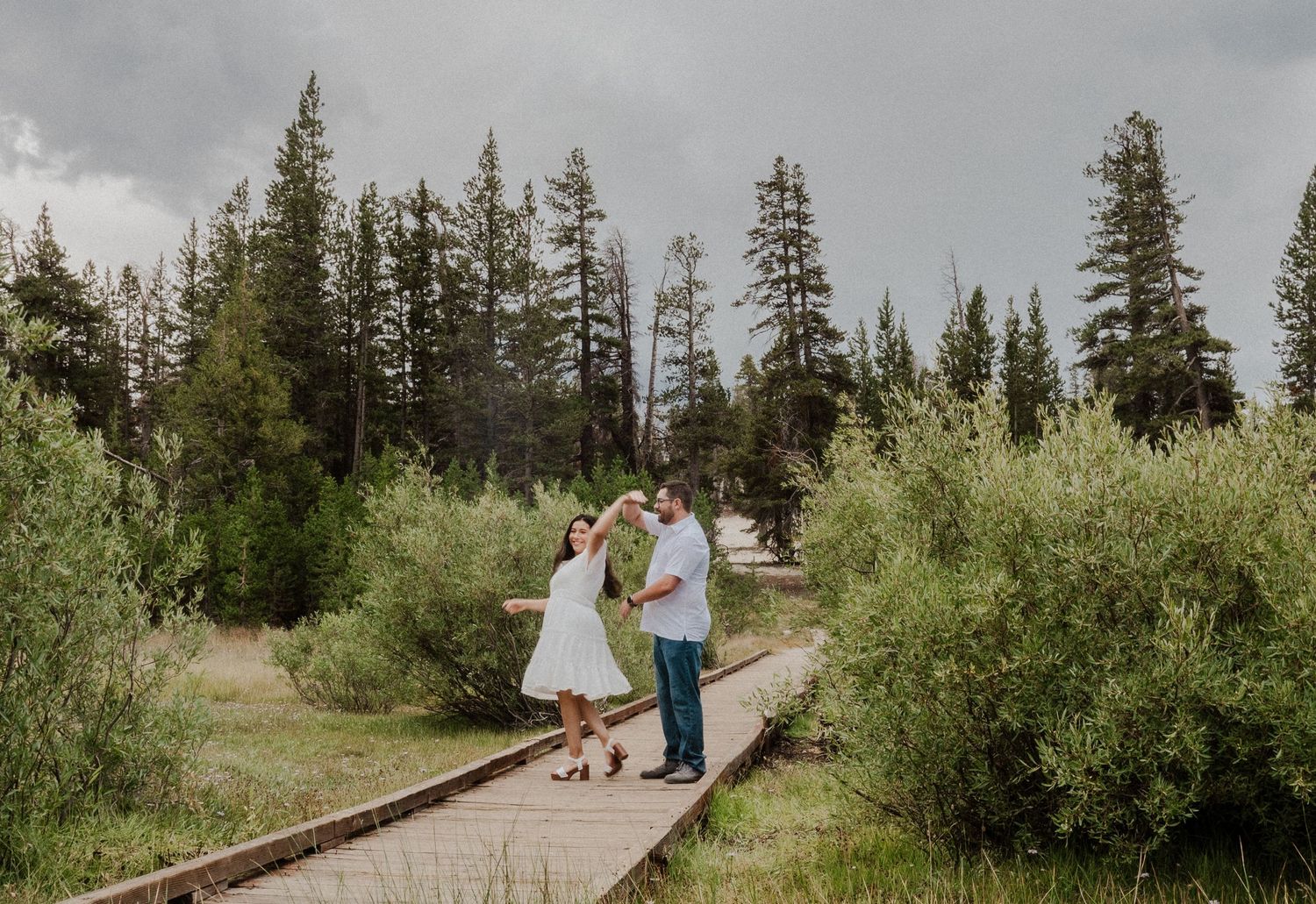 Couple dancing on a wooden boardwalk surrounded by lush green trees and shrubs under a cloudy sky.