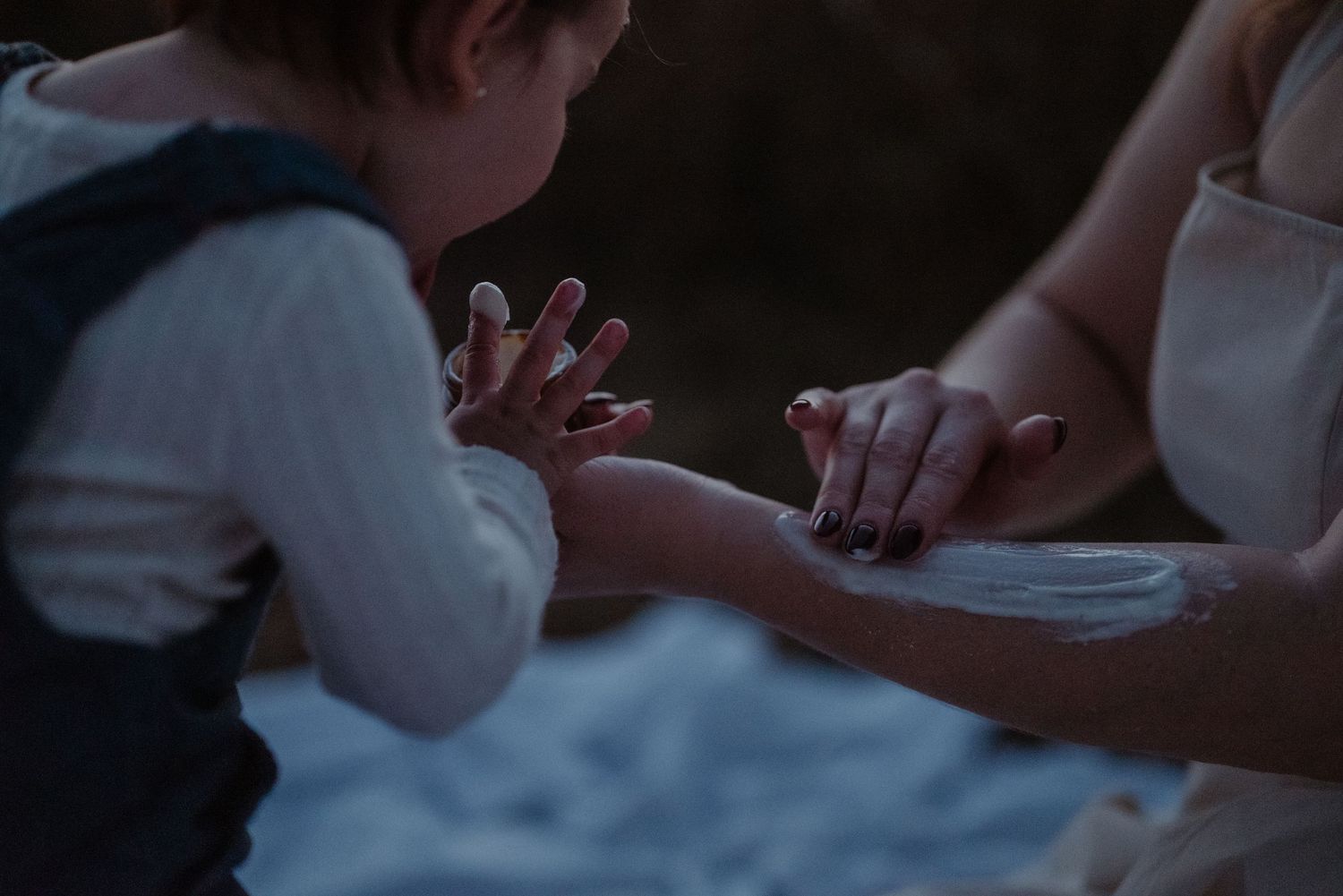 A child gently touches a woman's hand in a tender, intimate outdoor moment captured in soft, moody lighting.