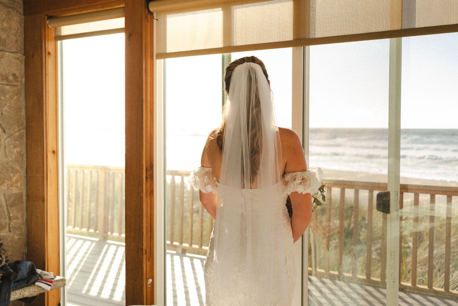 Bride in white wedding dress and veil stands gazing at ocean view through glass doors on sunny day.