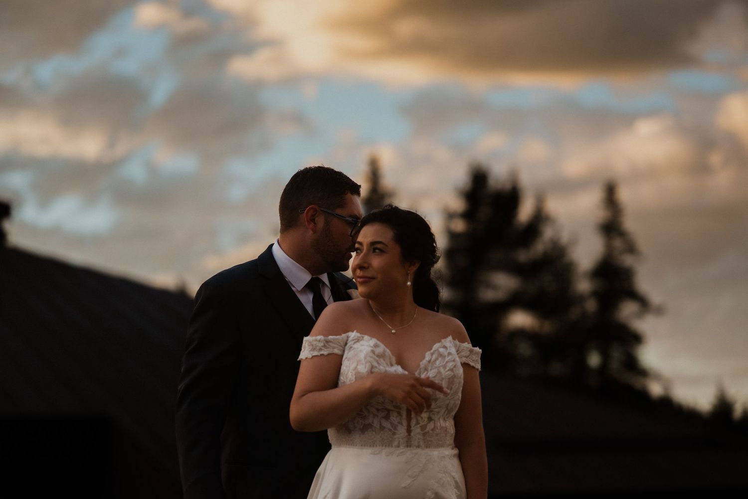 Bride and groom share a romantic moment at sunset with dramatic clouds and trees in the background.