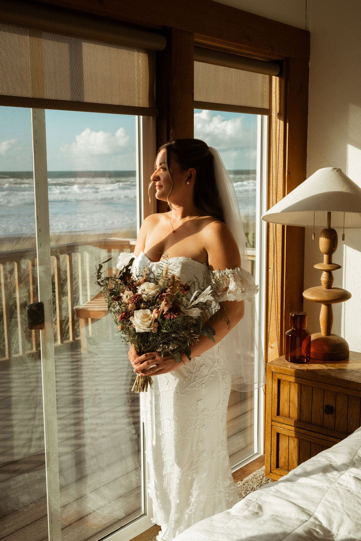 Bride in white lace off-shoulder gown holding floral bouquet, smiling by ocean-view window with veil.