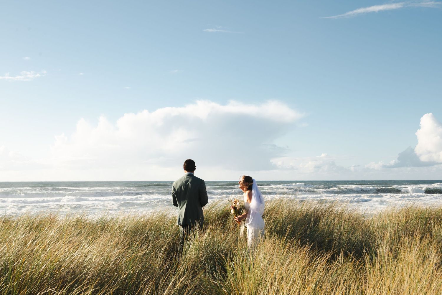 Bride and groom stand facing the ocean in tall beach grass, holding a bouquet under a bright blue sky with soft clouds.
