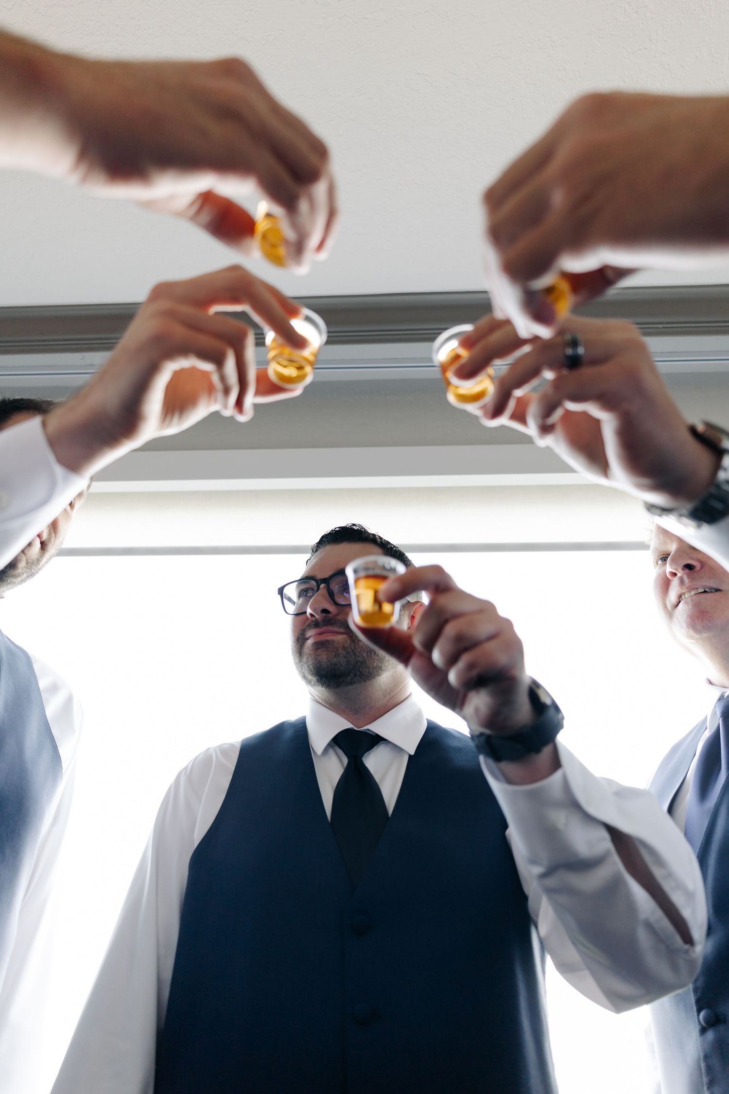 Groom in vest and tie toasts shots with groomsmen, low-angle view of hands raising glasses together indoors.