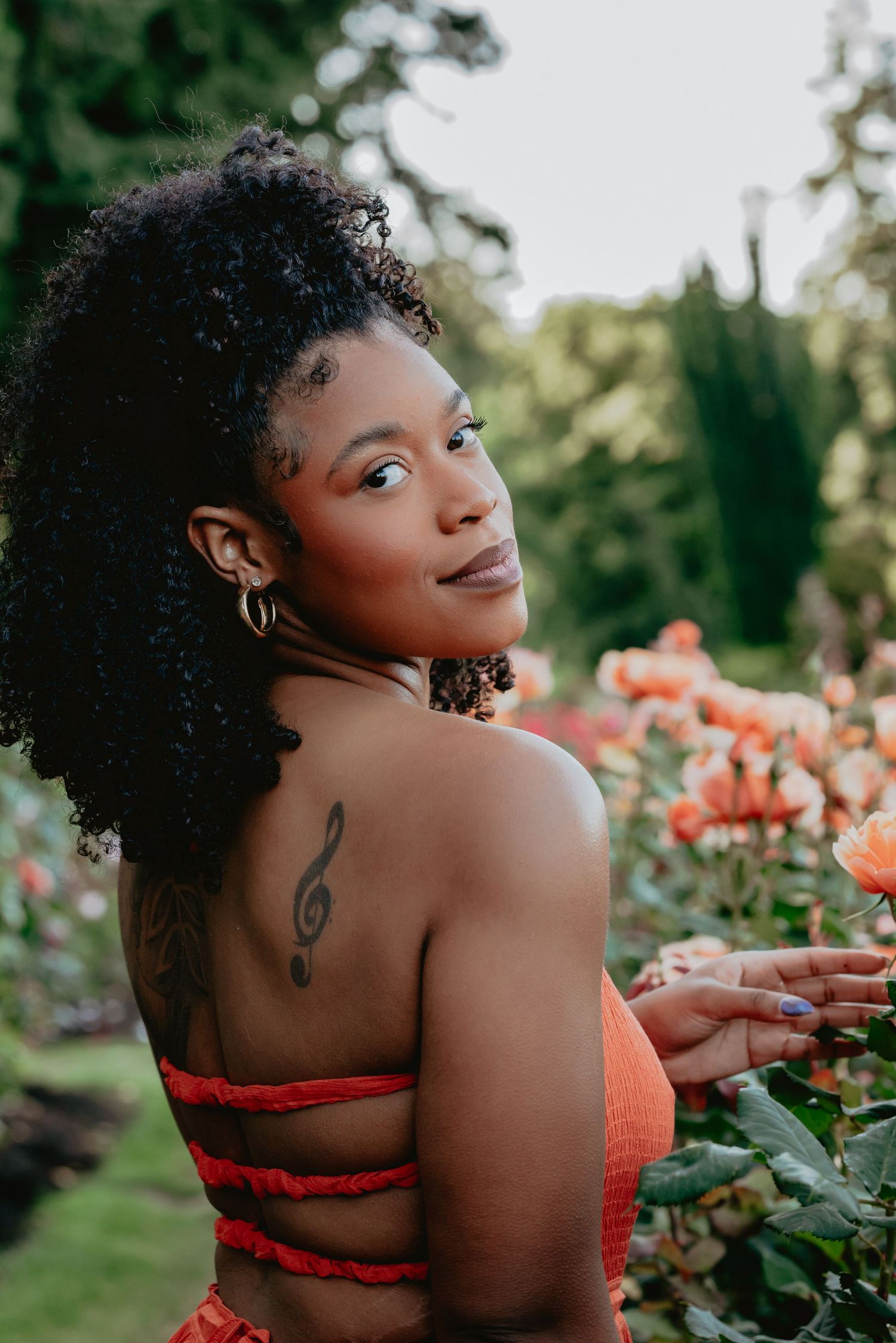 Young woman in orange top with treble clef tattoo poses among blooming roses in a lush garden.