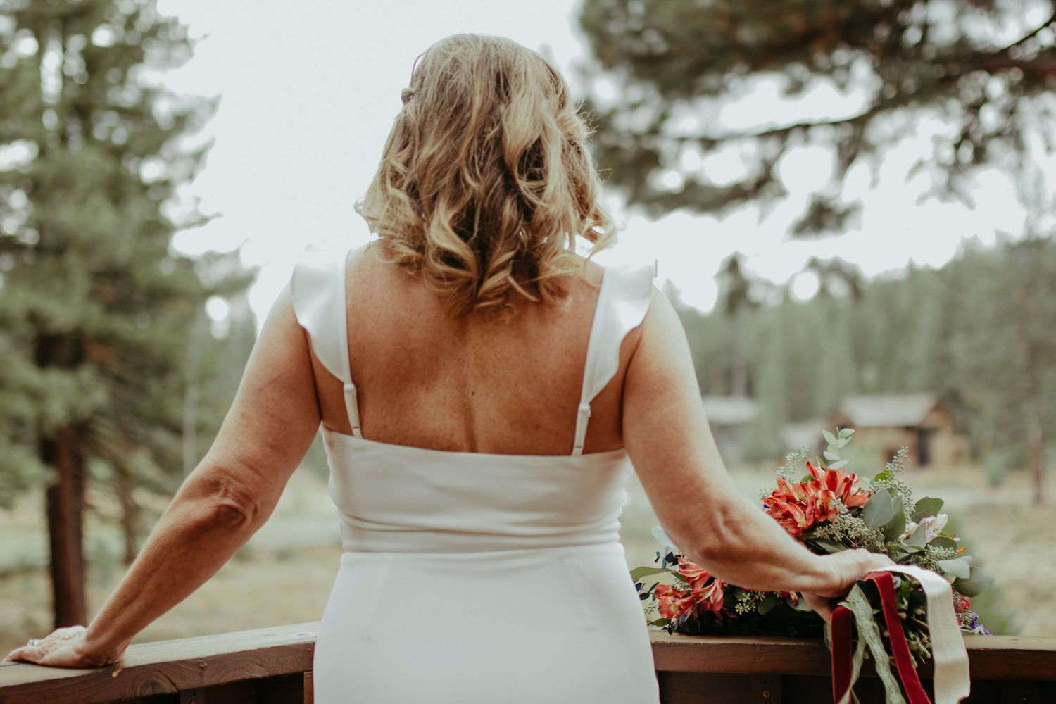 Bride in white wedding dress holding bouquet, viewed from behind on wooden deck surrounded by pine trees.