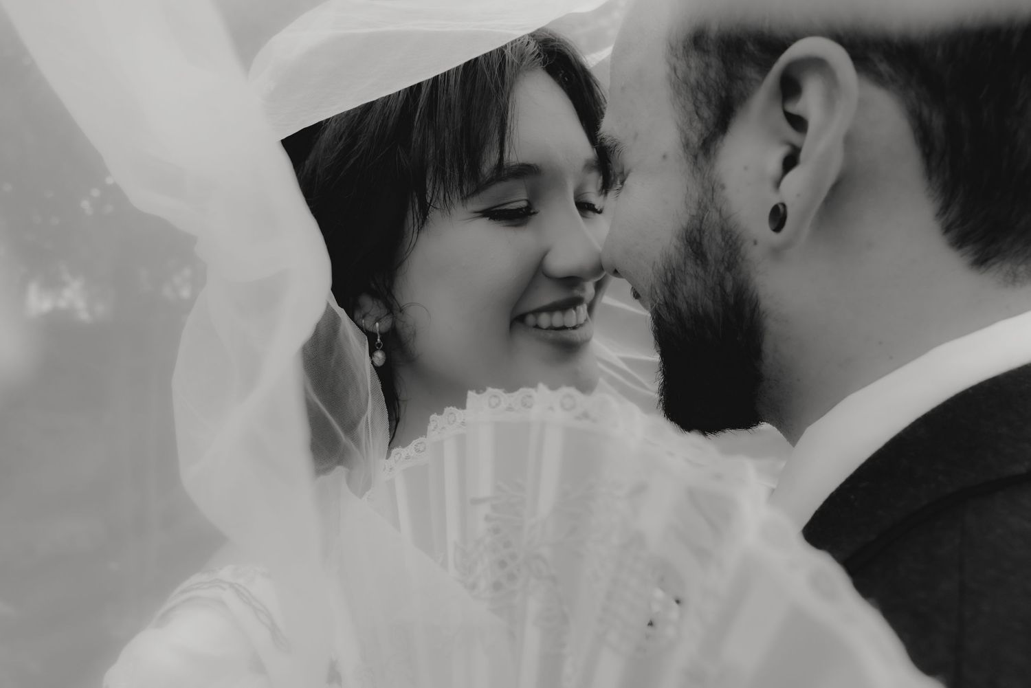 Black and white wedding photo of smiling bride and groom nearly kissing under a flowing veil.