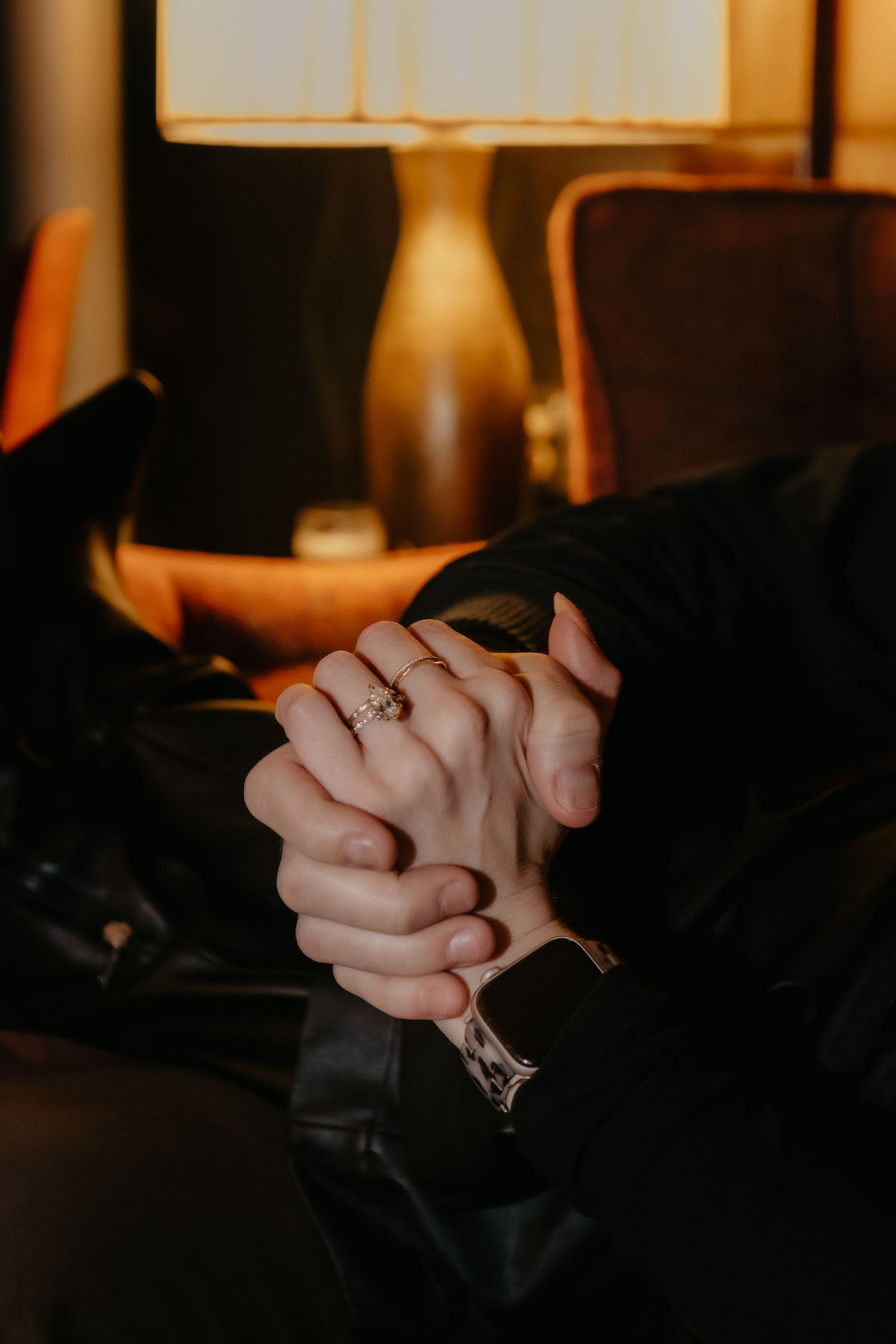Clasped hands with a gold ring in warm lamplight, wearing black clothing, seated in an orange chair.