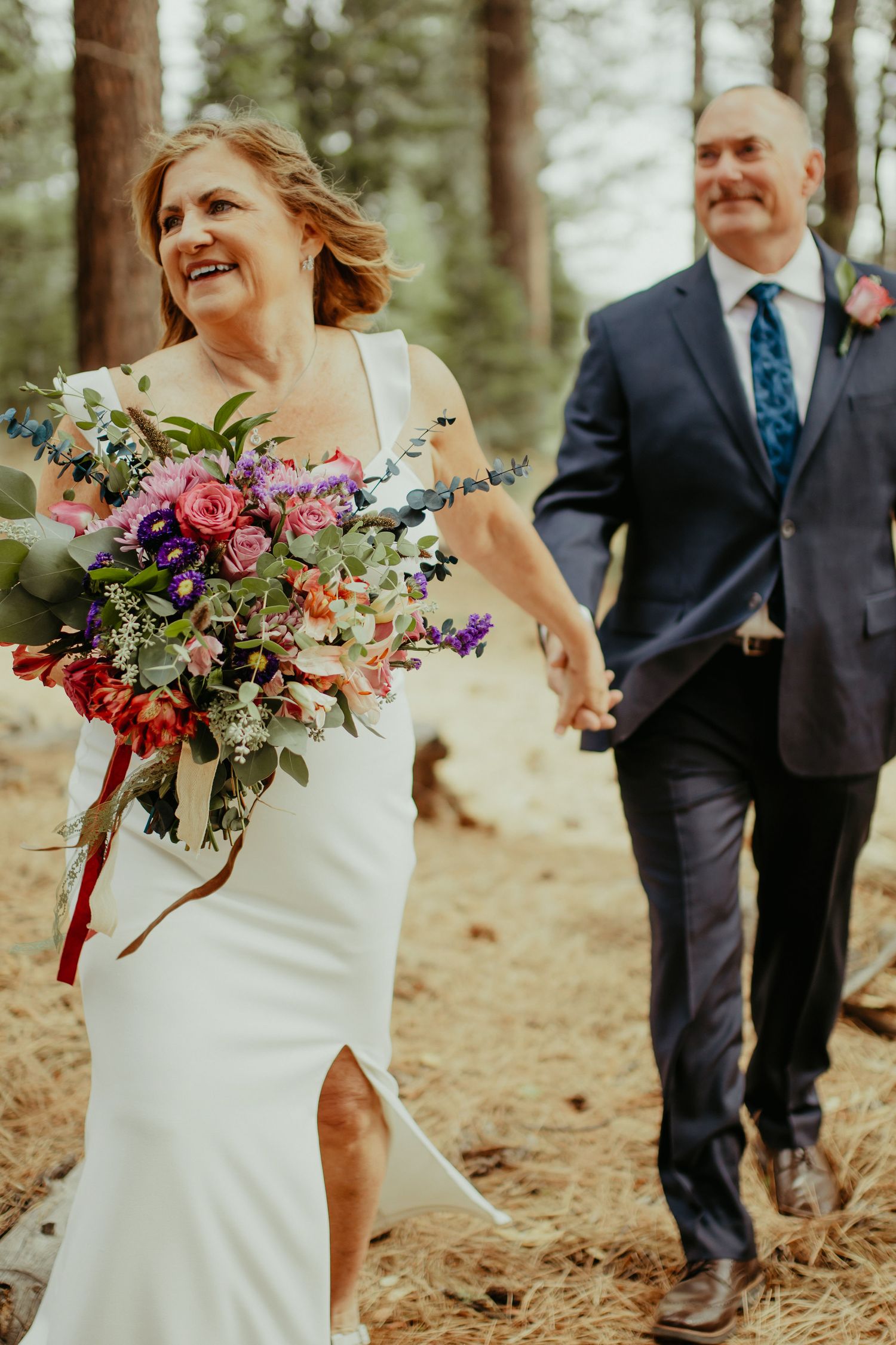 A joyful bride in a white gown holds a colorful bouquet while walking hand-in-hand with a groom in a navy suit outdoors.
