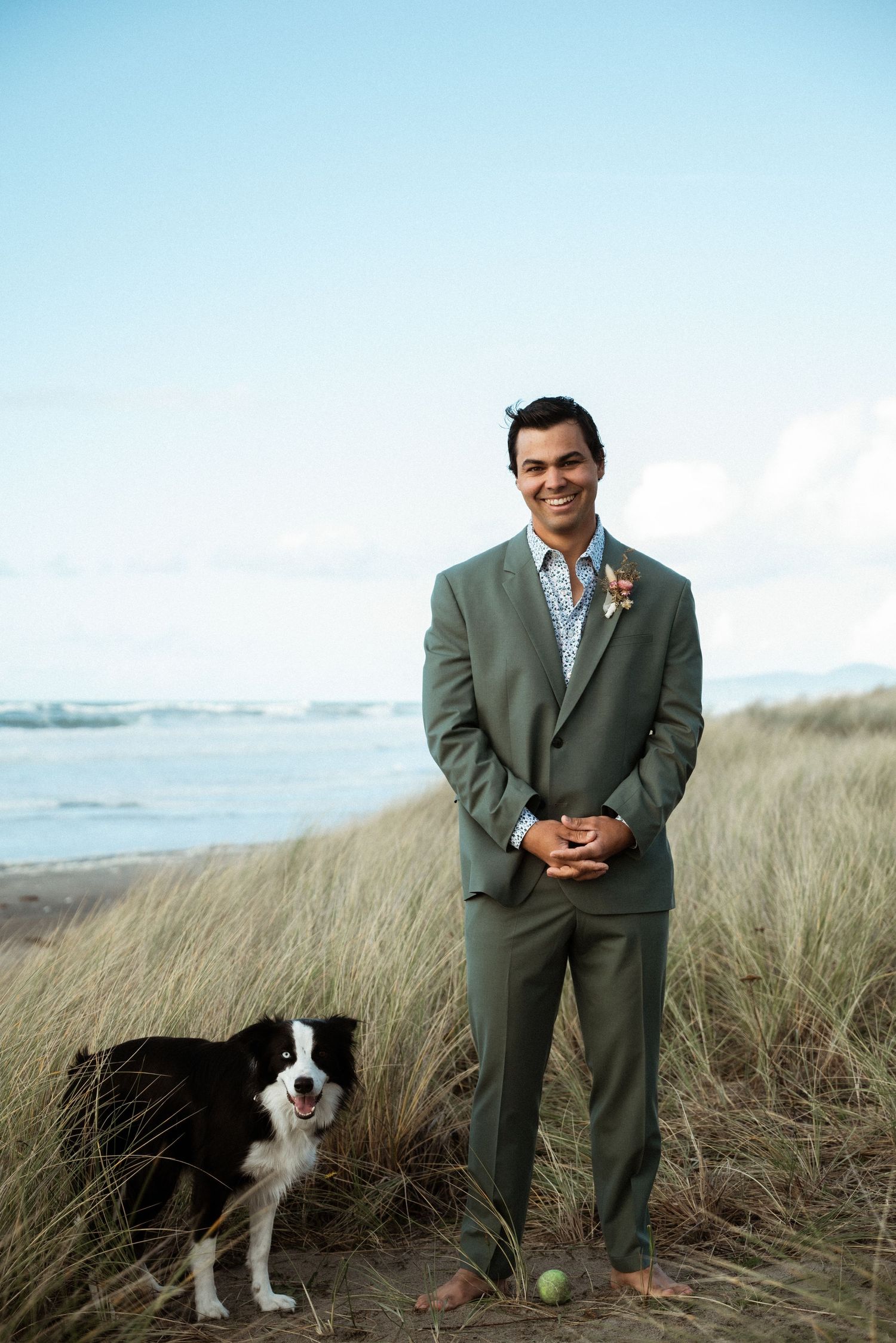Barefoot groom in an olive green suit stands in beach dune grass beside a border collie with a tennis ball at his feet.