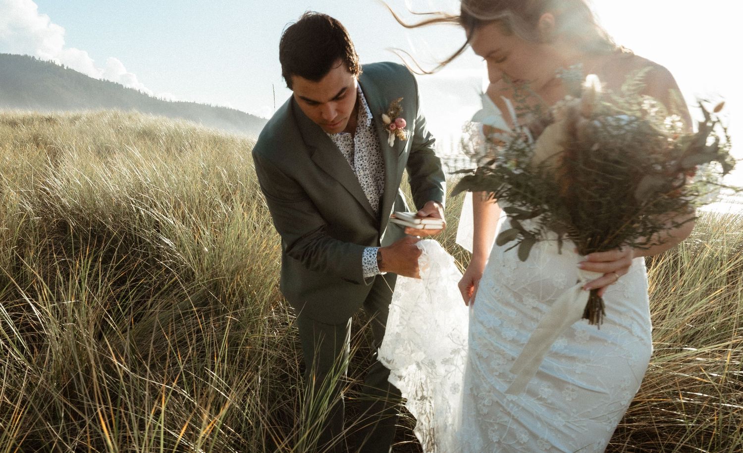 A groom in a gray suit leans toward a bride in a white gown holding a boho bouquet in windswept coastal dune grass.