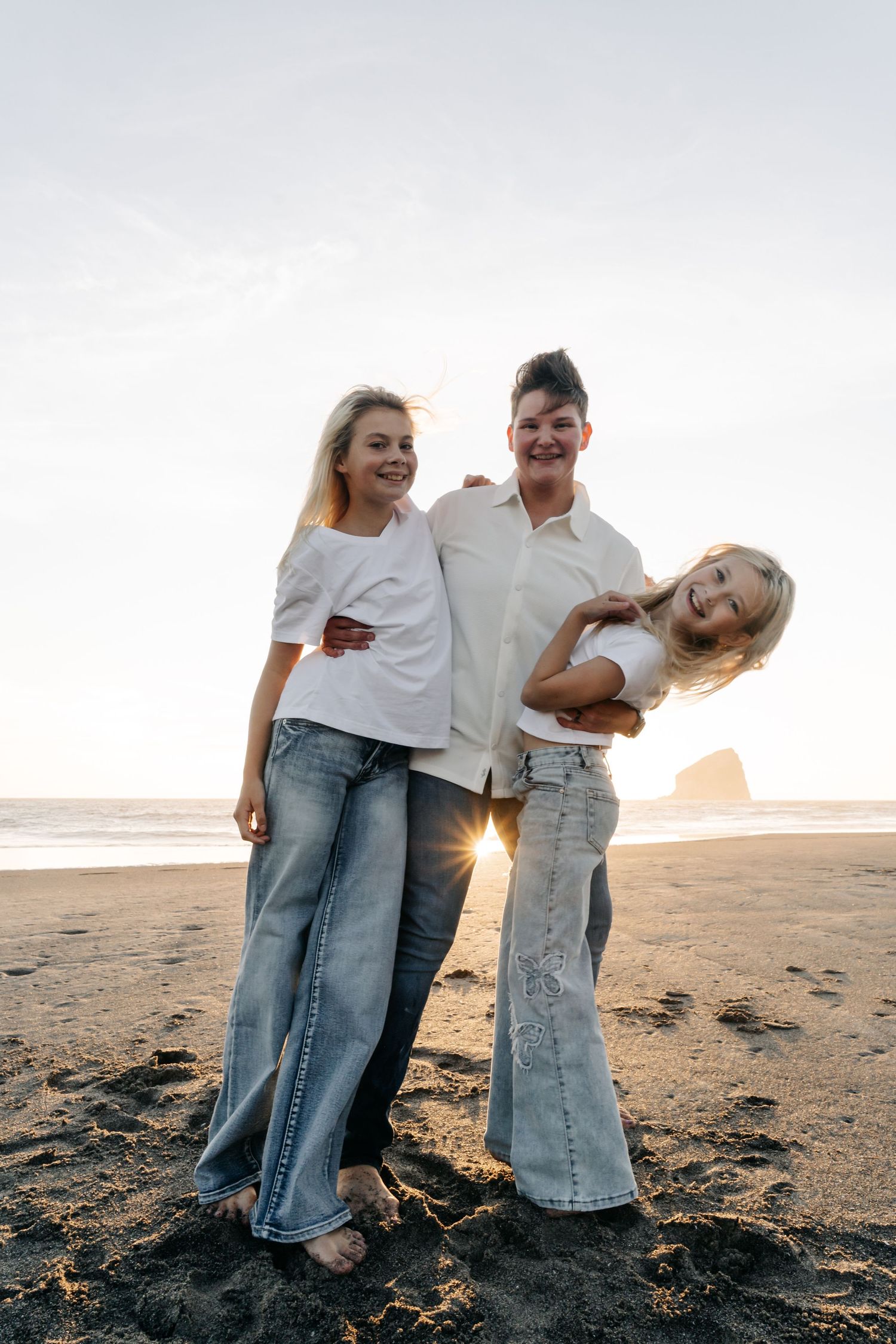 Three women in white tops and jeans laughing together on a sandy beach at sunset.