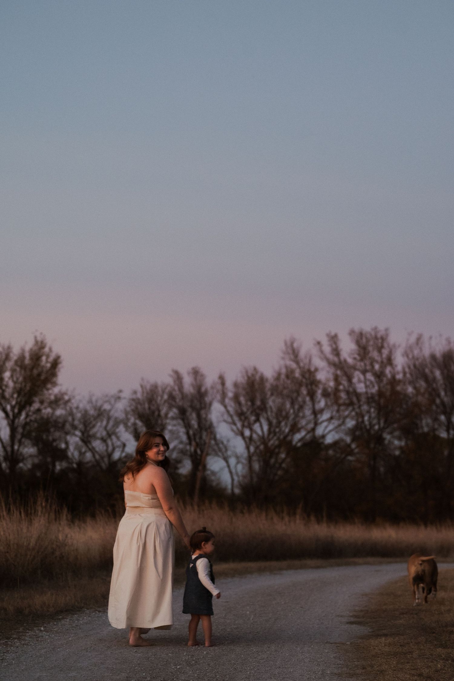 Woman and child walk hand-in-hand on a rural path at dusk with trees and grazing animals nearby.