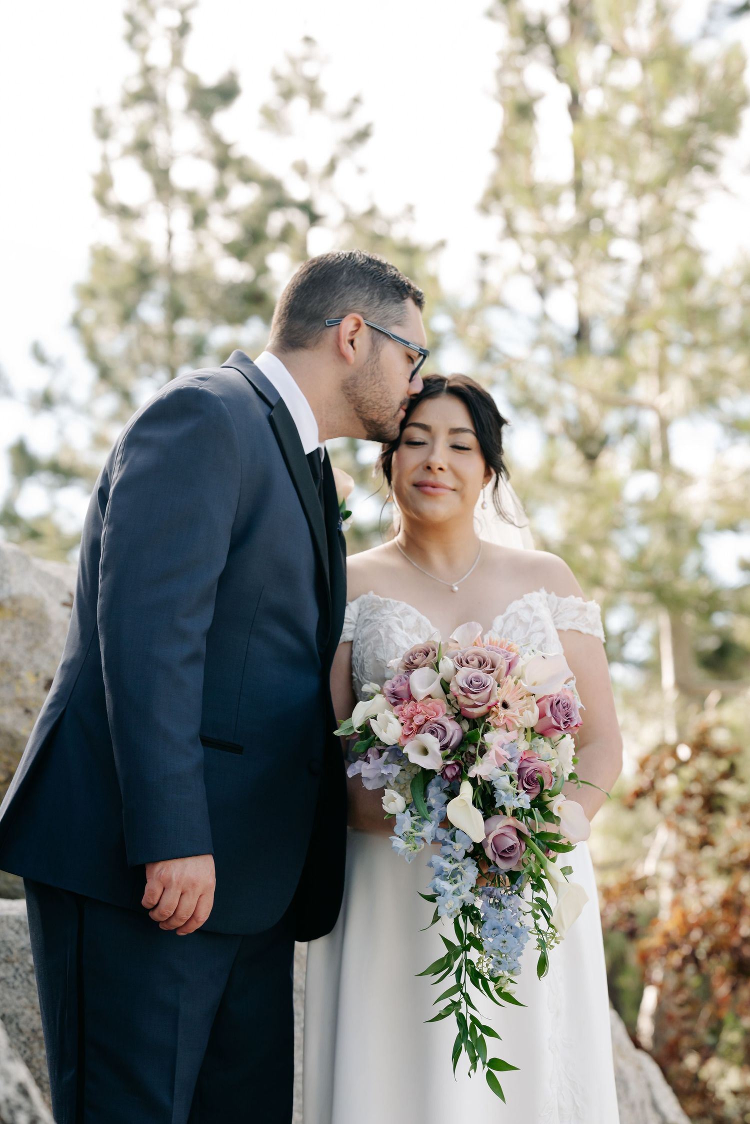 Groom in navy suit kissing bride's cheek as she holds cascading pink and blue floral bouquet outdoors.