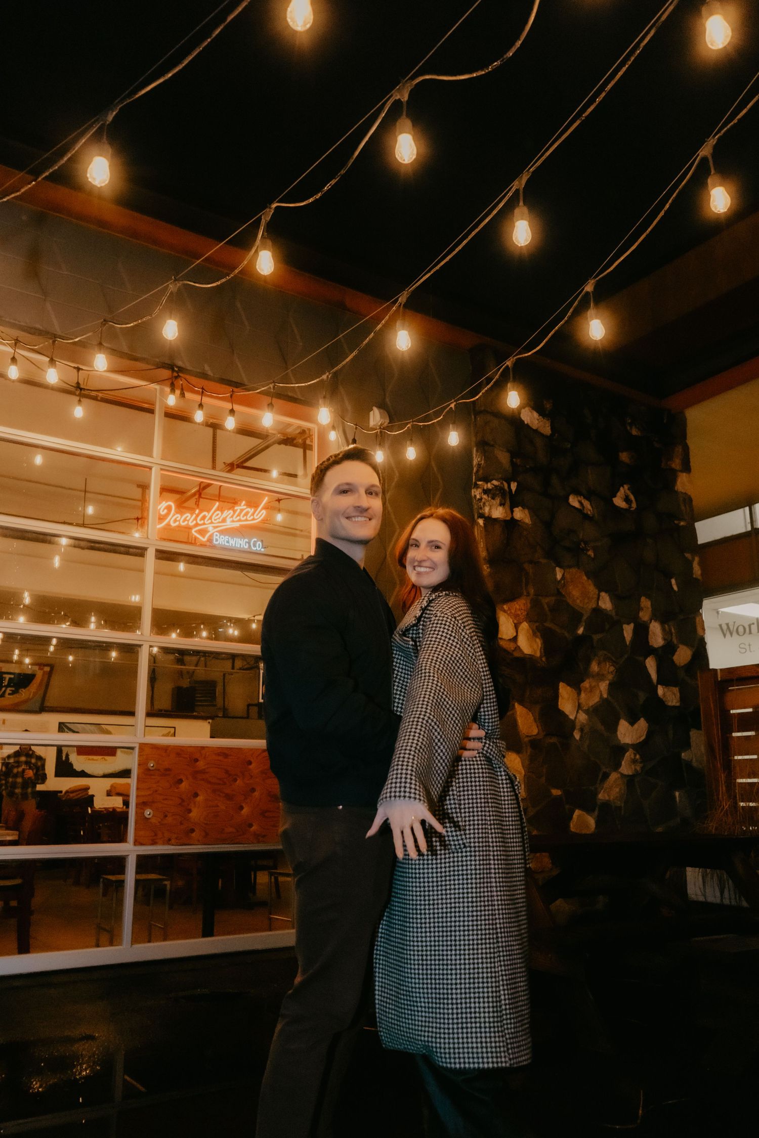 Couple smiling together under warm string lights outside a cozy restaurant with stone wall decor at night.