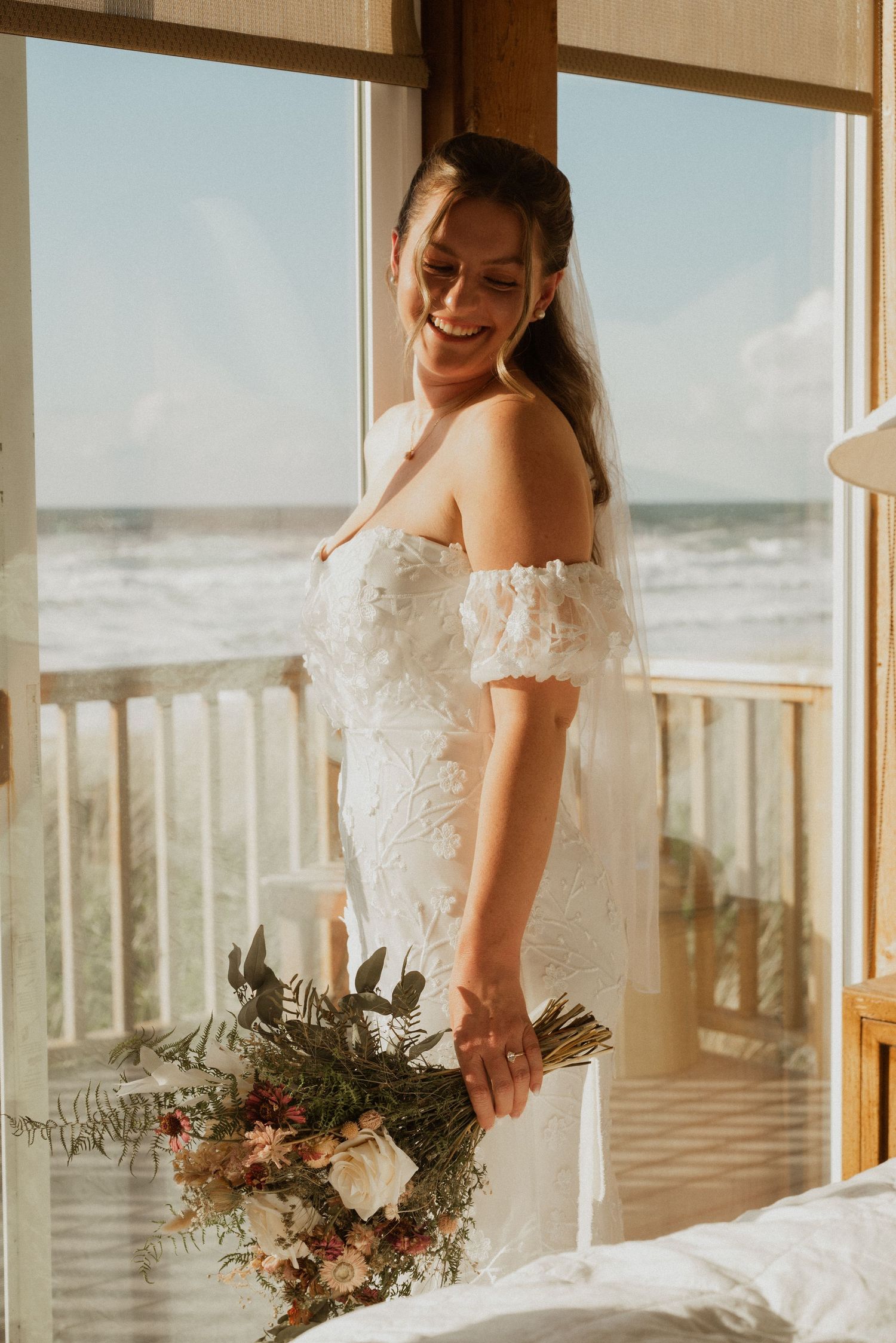 Smiling bride in an off-shoulder lace wedding gown holds a rustic bouquet beside a window with an ocean view.