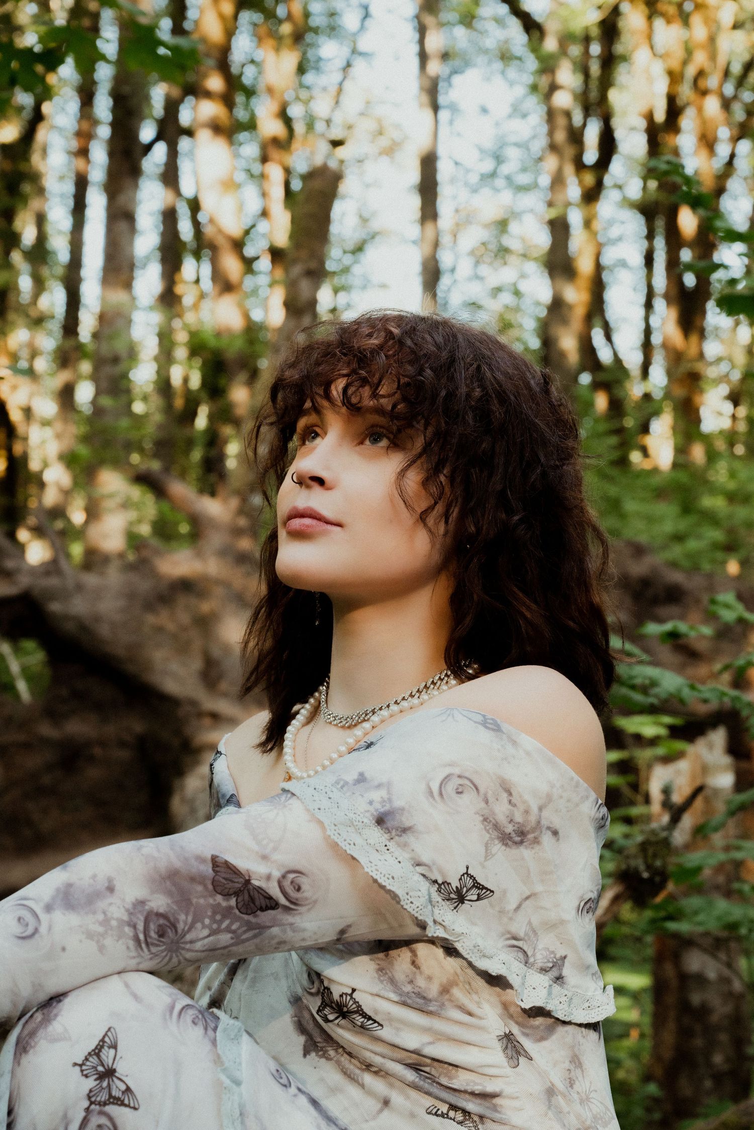 Woman in floral off-shoulder dress with pearl necklace sitting in a sunlit forest, gazing upward.