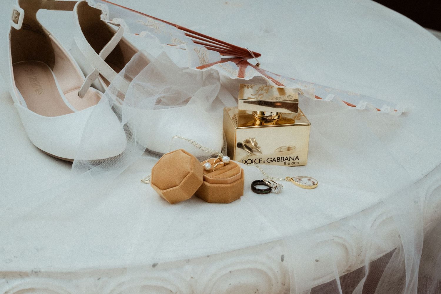 White bridal heels, Dolce & Gabbana perfume, gold ring box, and elegant jewelry arranged on a white surface.