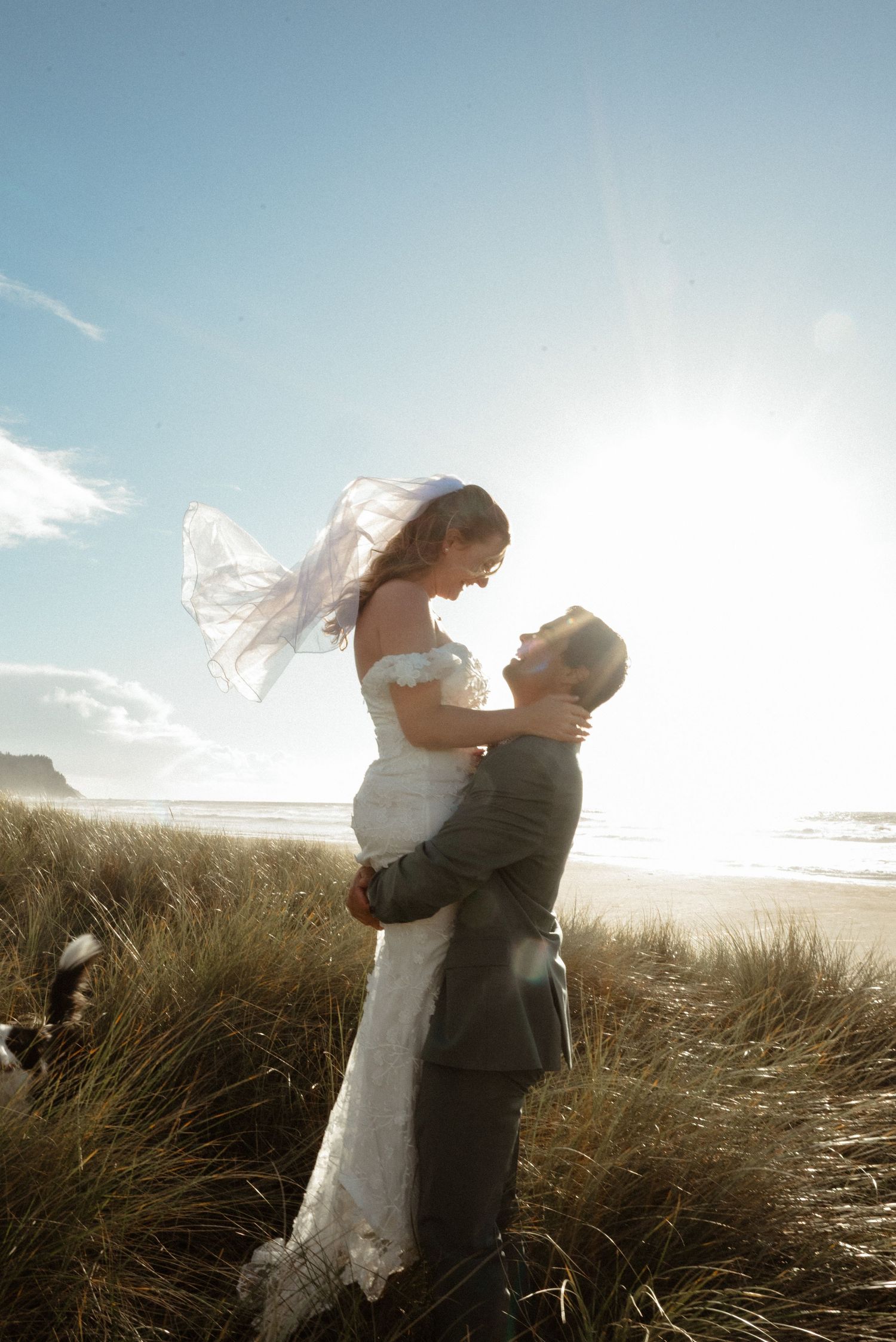 Groom lifts his bride in a flowing white gown and veil as they embrace among beach dune grass at sunset by the ocean.